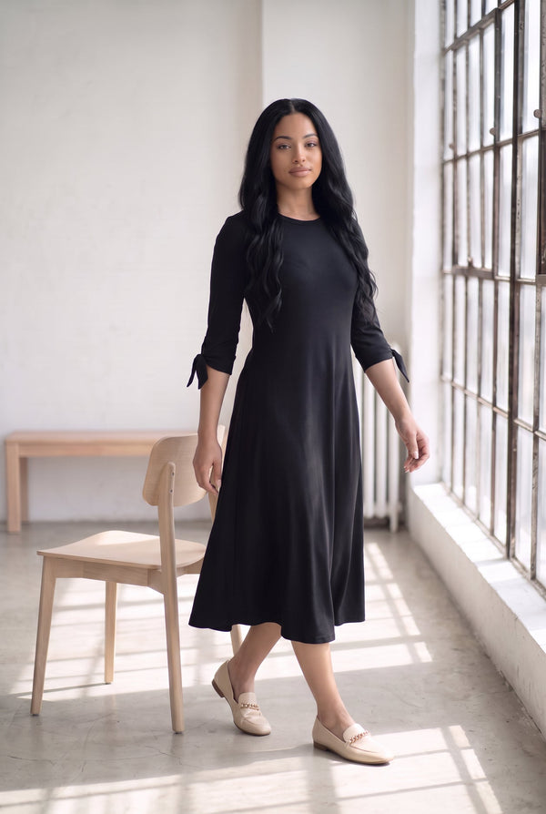 Nuggles® Gracie Dress in Black, full length mid-stride by a sunny window in cream loafers
