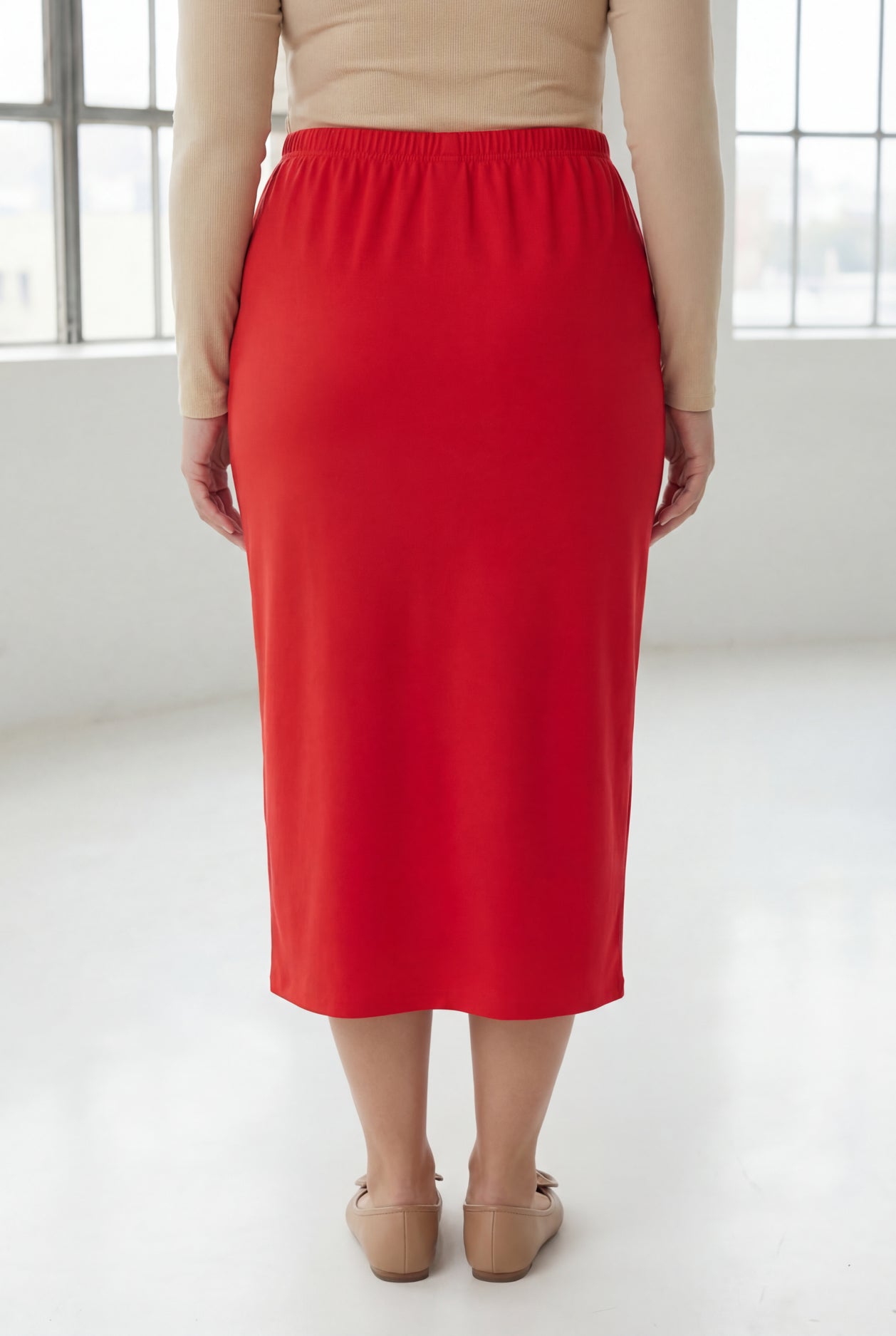 Woman wearing a beige top and red skirt in a bright room with large windows.