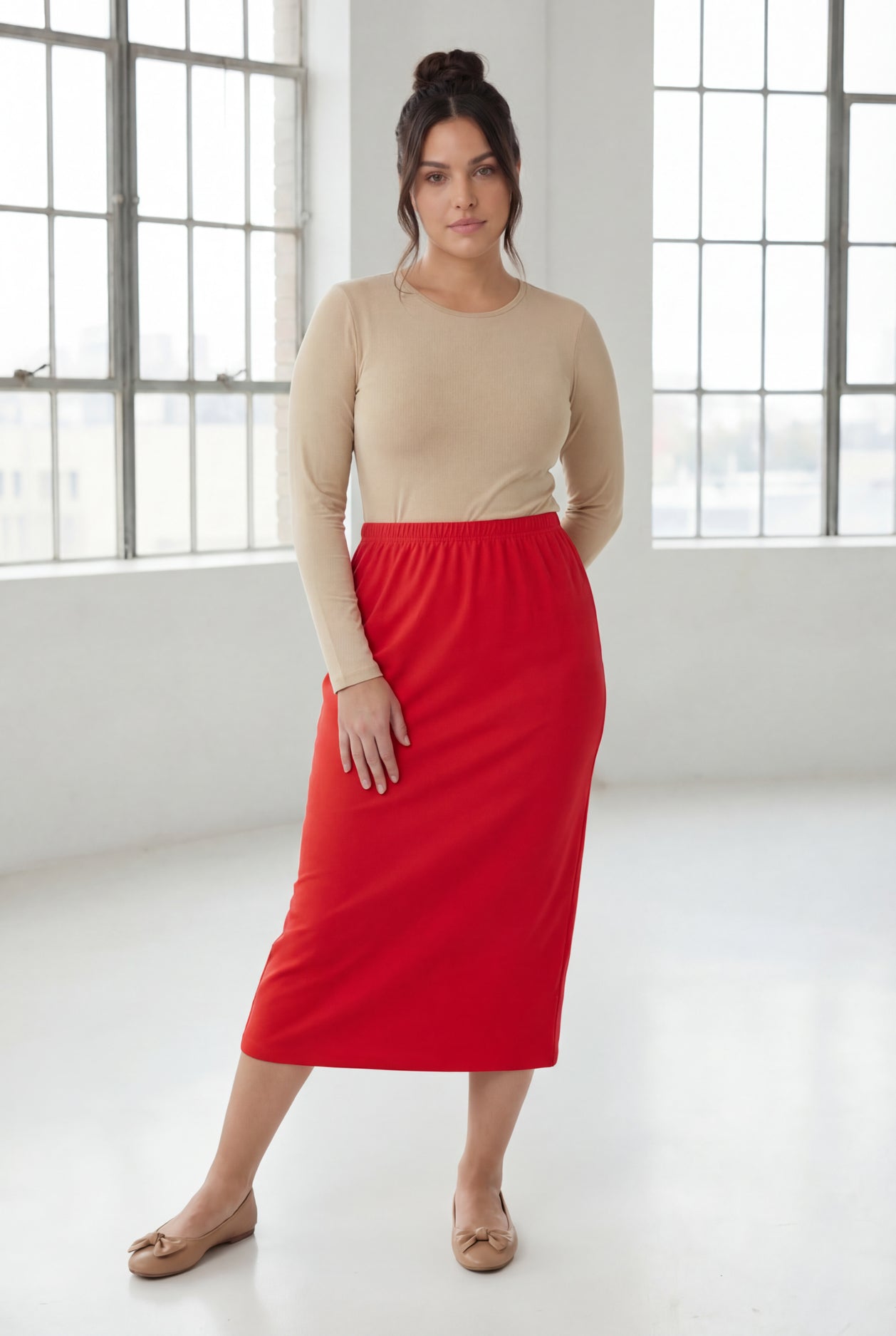 Woman wearing a beige top and red skirt in a bright room with large windows.