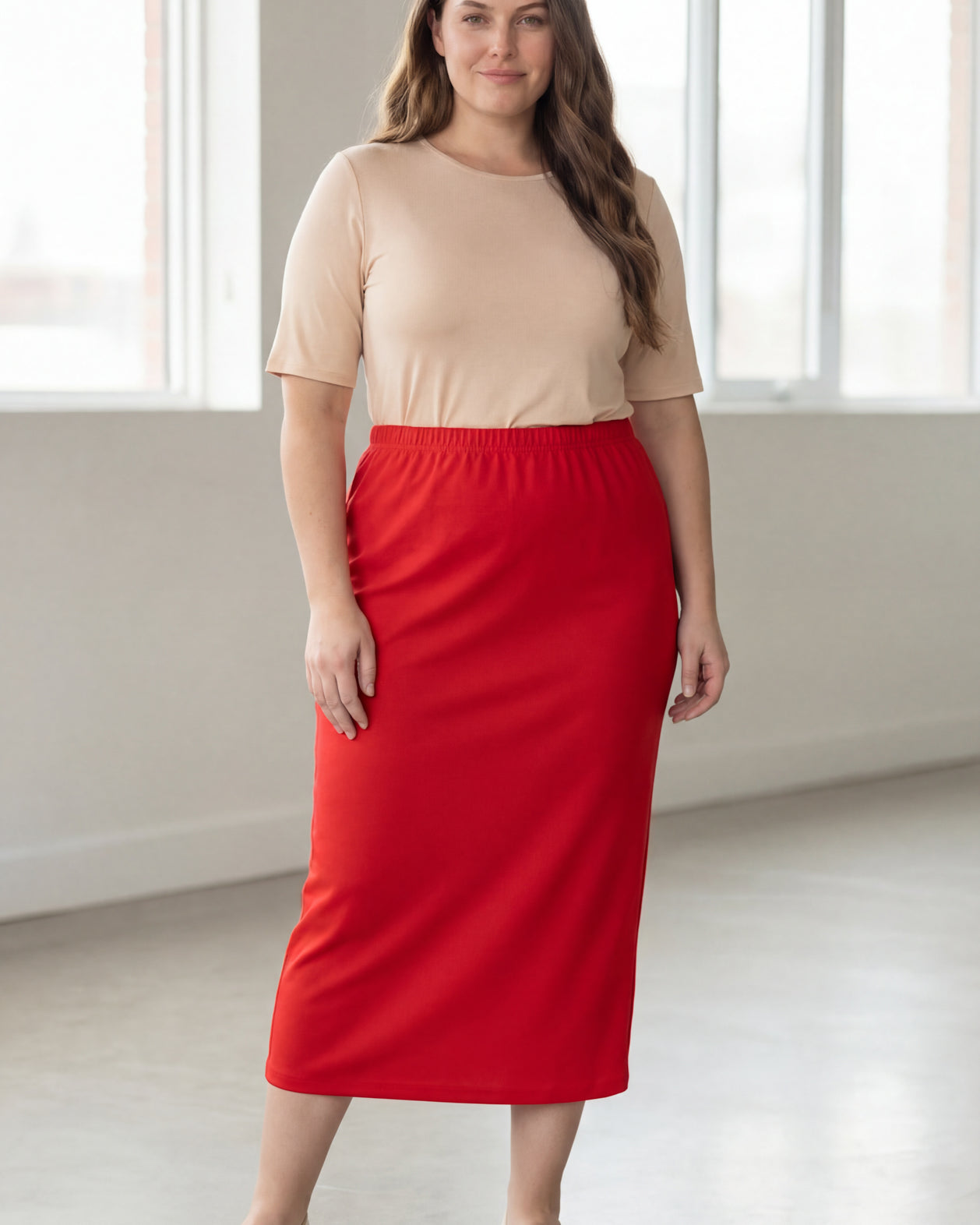 Woman wearing a beige shirt & red maxi length pencil skirt standing in a bright room with large windows.