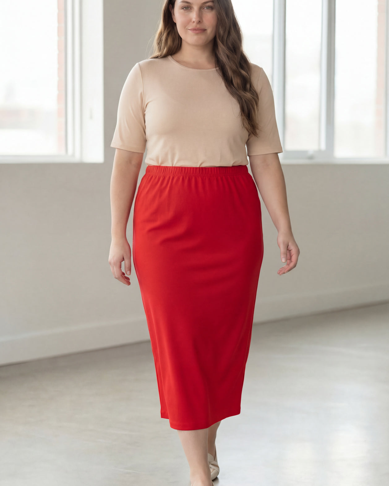 Woman wearing a beige shirt & red maxi length pencil skirt standing in a bright room with large windows.