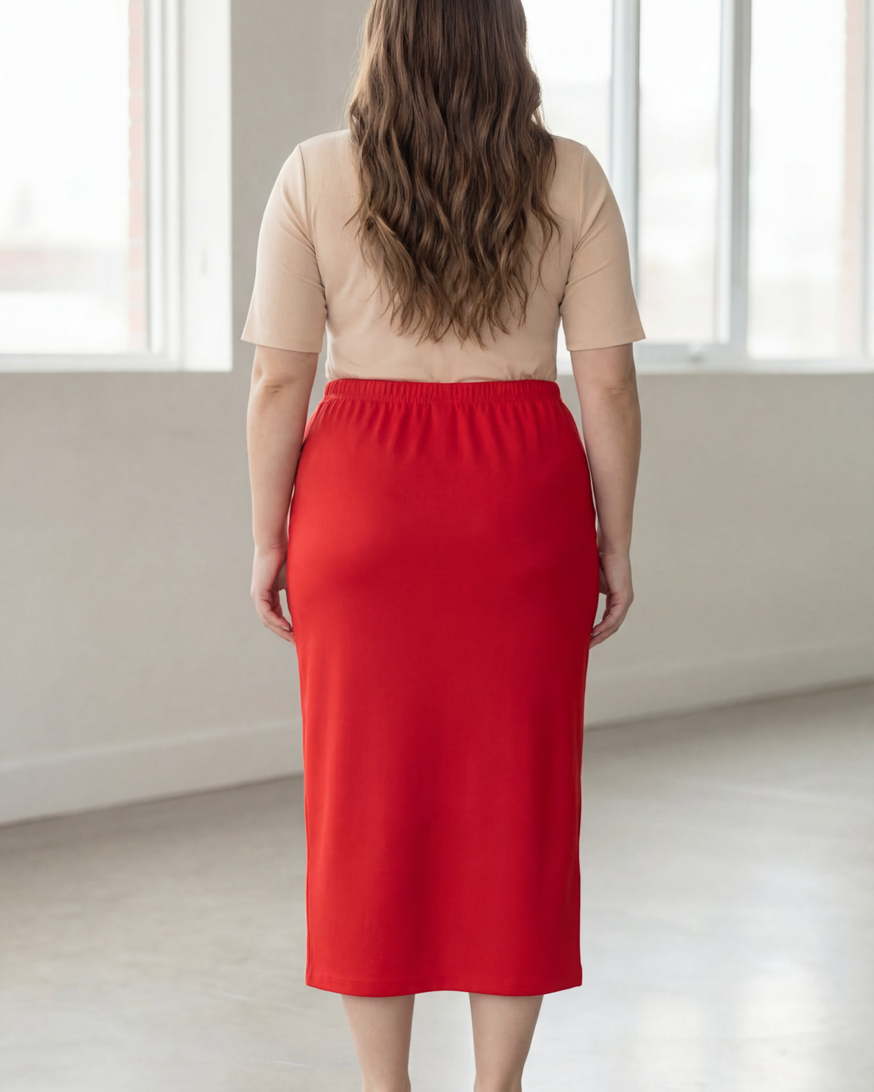 Woman wearing a beige shirt & red maxi length pencil skirt standing in a bright room with large windows.