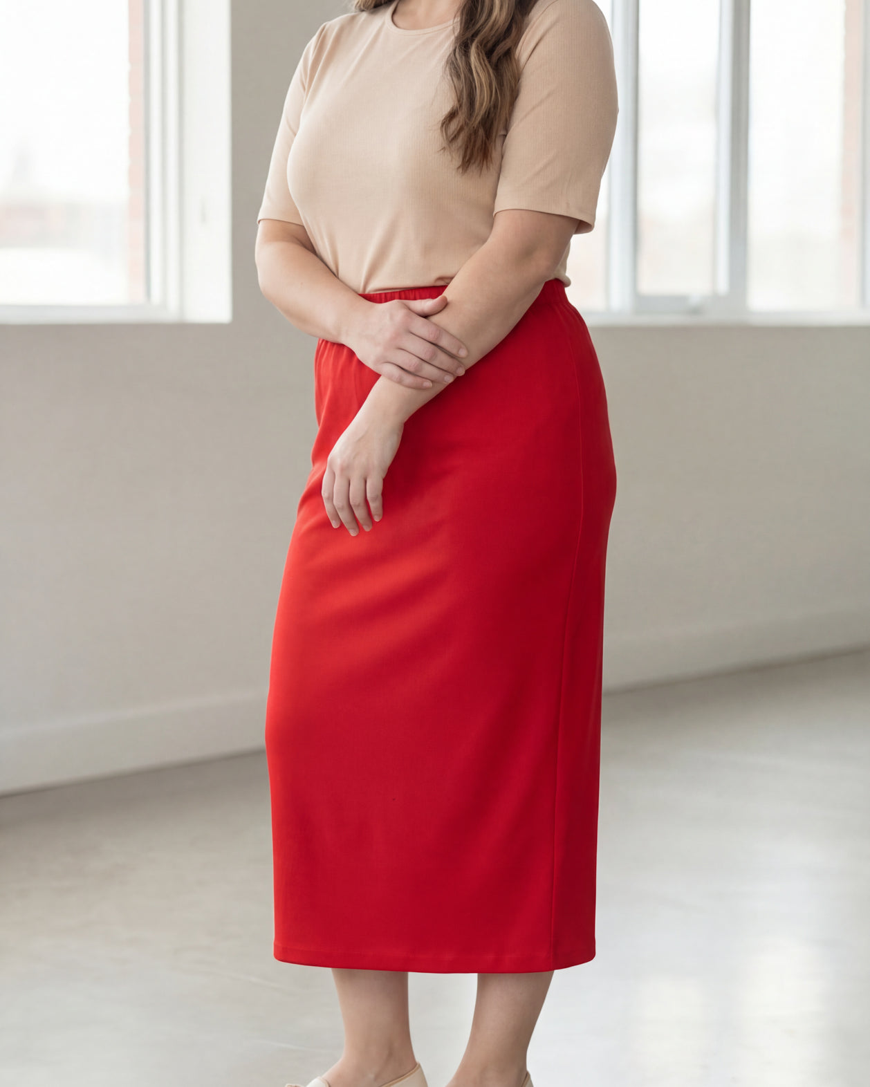 Woman wearing a beige shirt & red maxi length pencil skirt standing in a bright room with large windows.