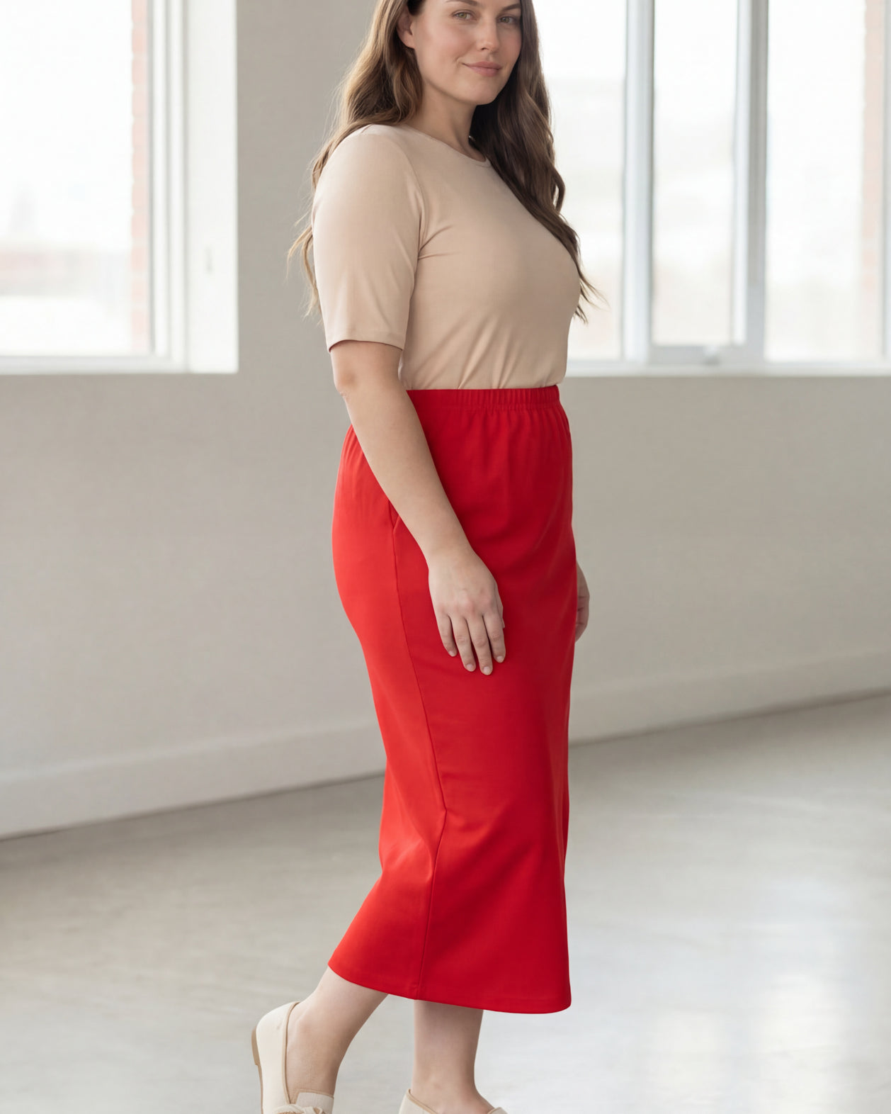 Woman wearing a beige shirt & red maxi length pencil skirt standing in a bright room with large windows.
