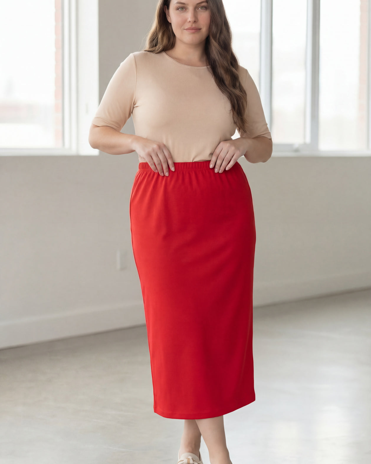 Woman wearing a beige shirt & red maxi length pencil skirt standing in a bright room with large windows.