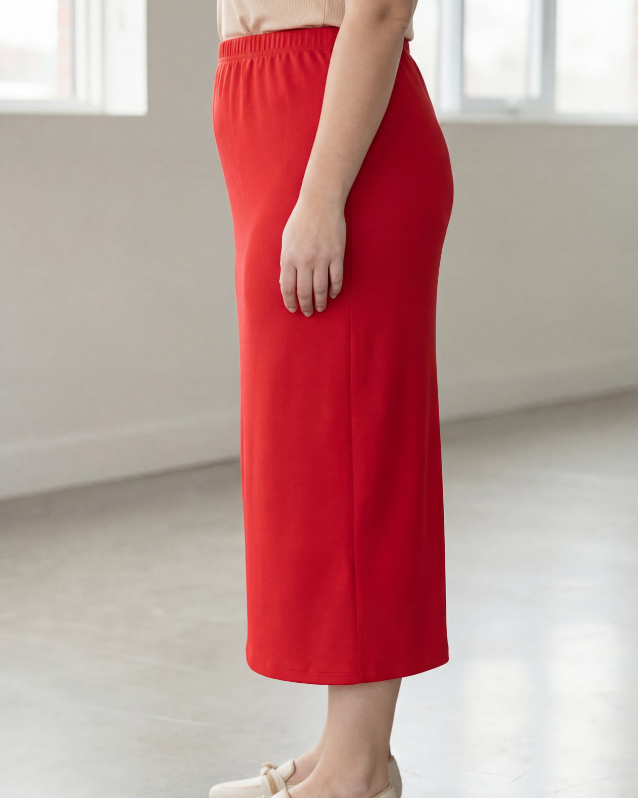 Woman wearing a beige shirt & red maxi length pencil skirt standing in a bright room with large windows.