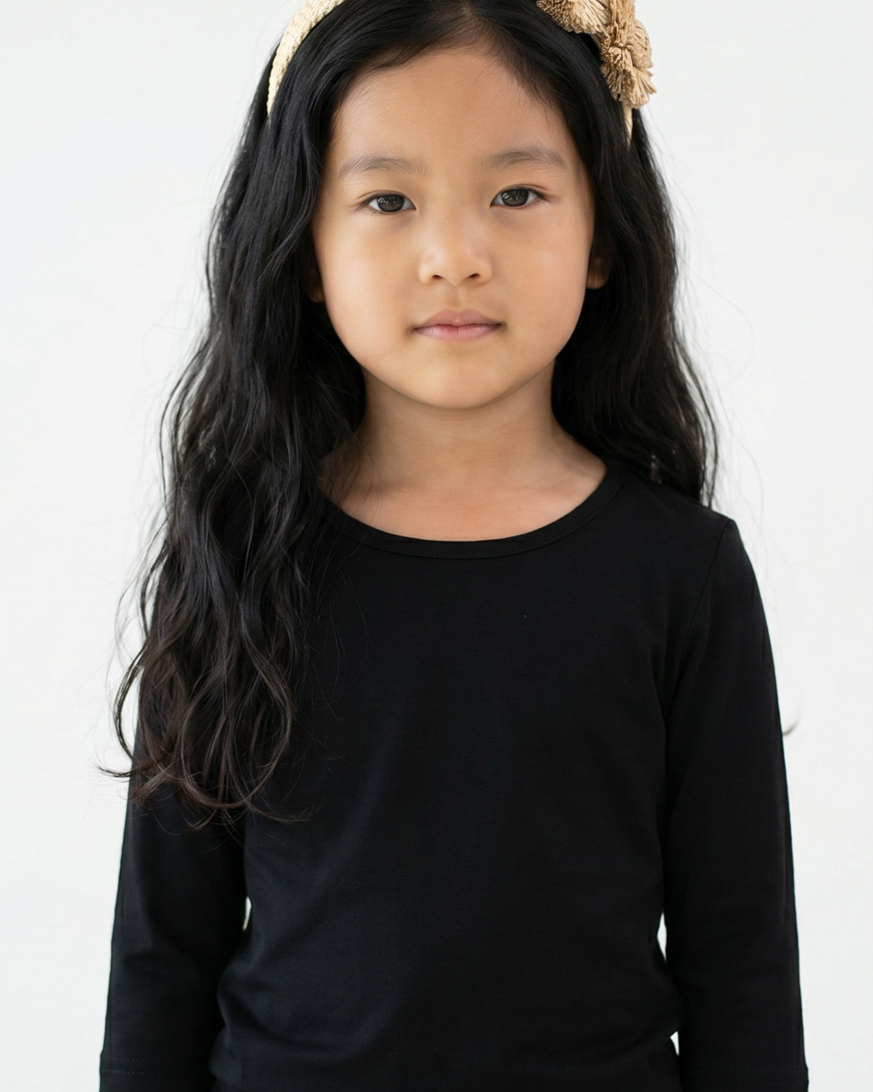 Young girl with long dark hair wearing Nuggles® Black Tee and a beige floral headband against a white background.