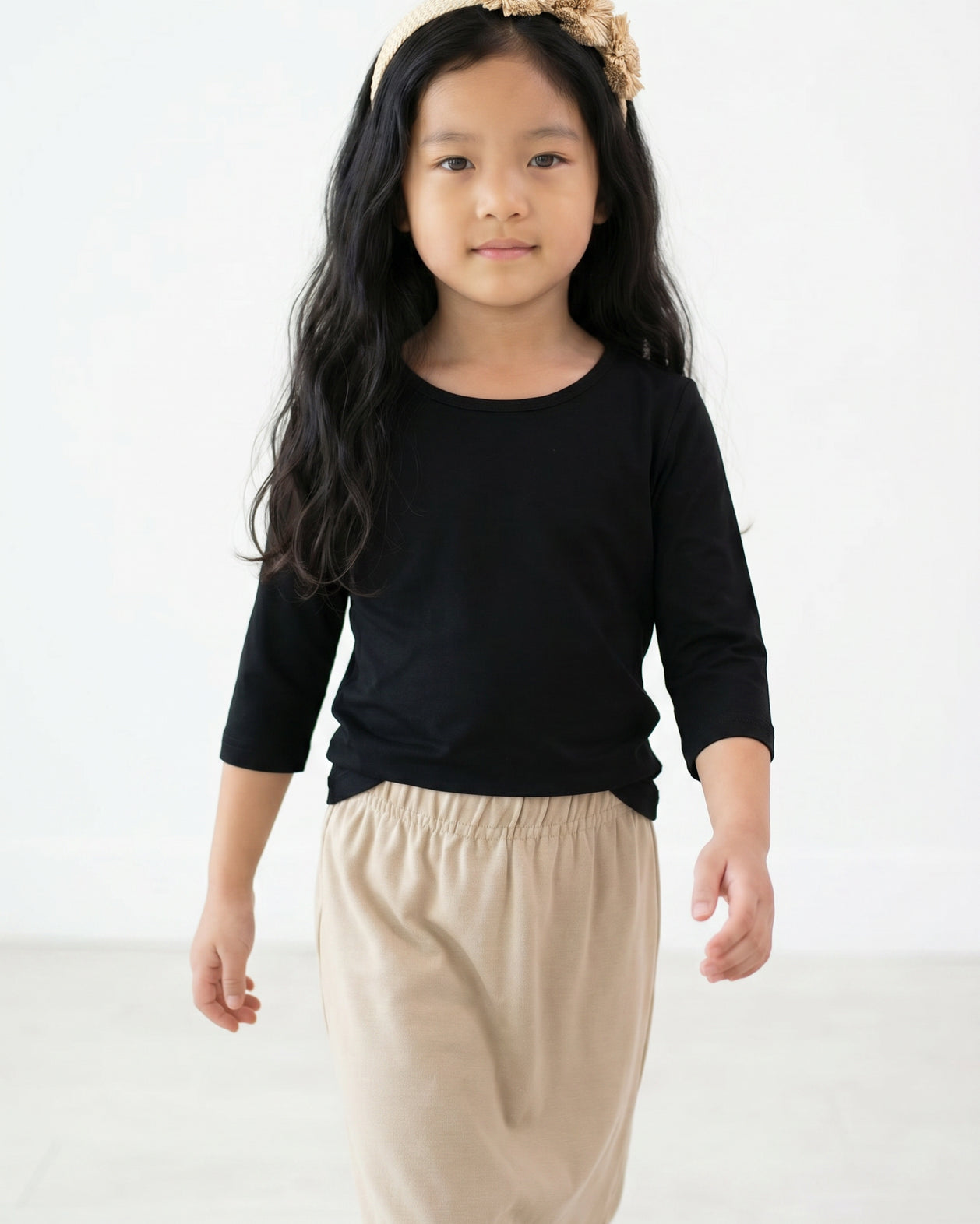 Young girl with long dark hair wearing Nuggles® Black Tee and a beige floral headband against a white background.