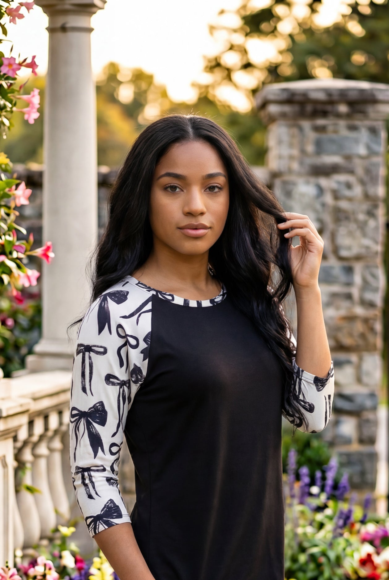 Person wearing a black and white patterned top standing in a garden with flowers and stone pillars.
