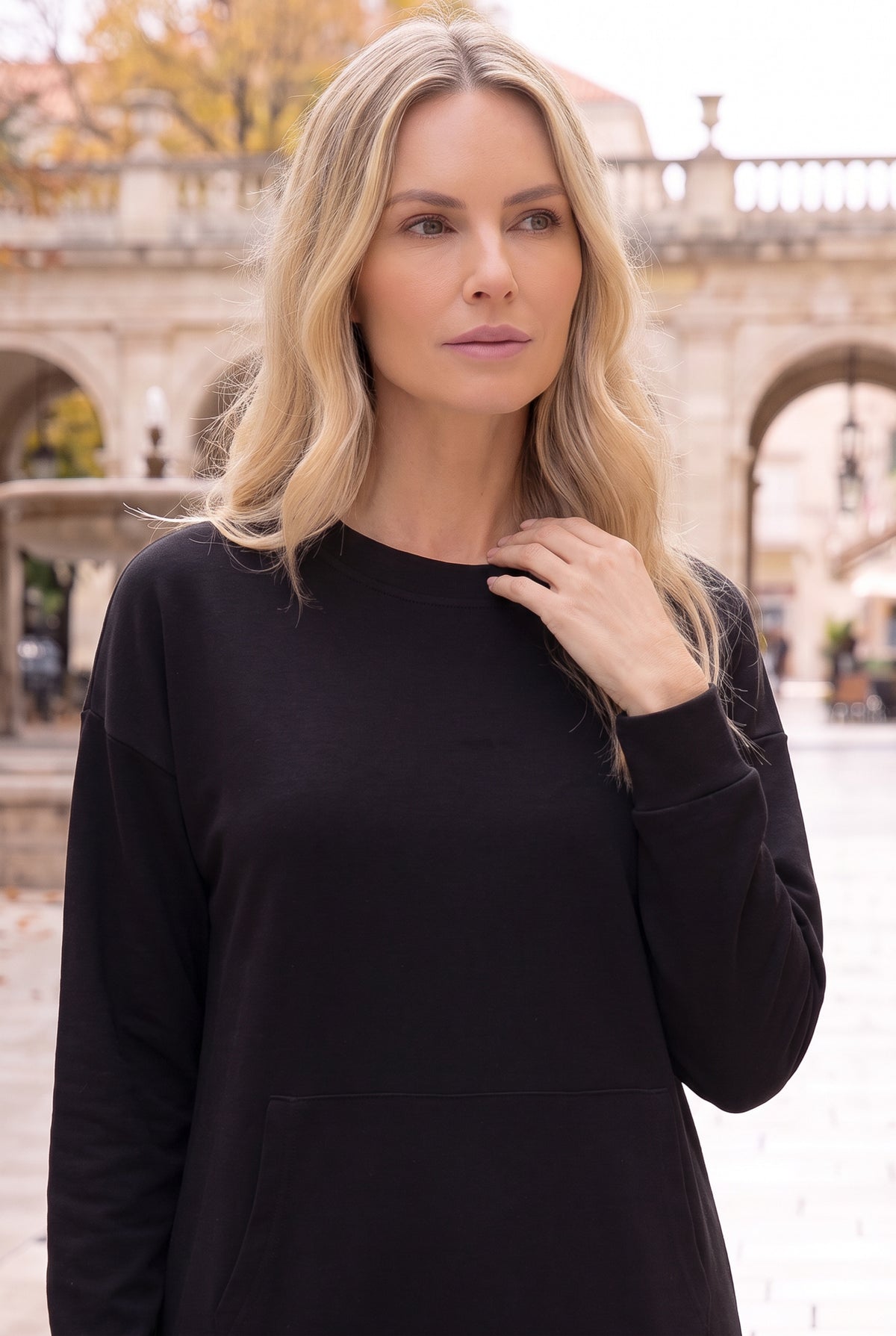 Nuggles® Oakley Hoodie Dress in Black, waist-up view with a hand at the ribbed crew neckline in a stone courtyard