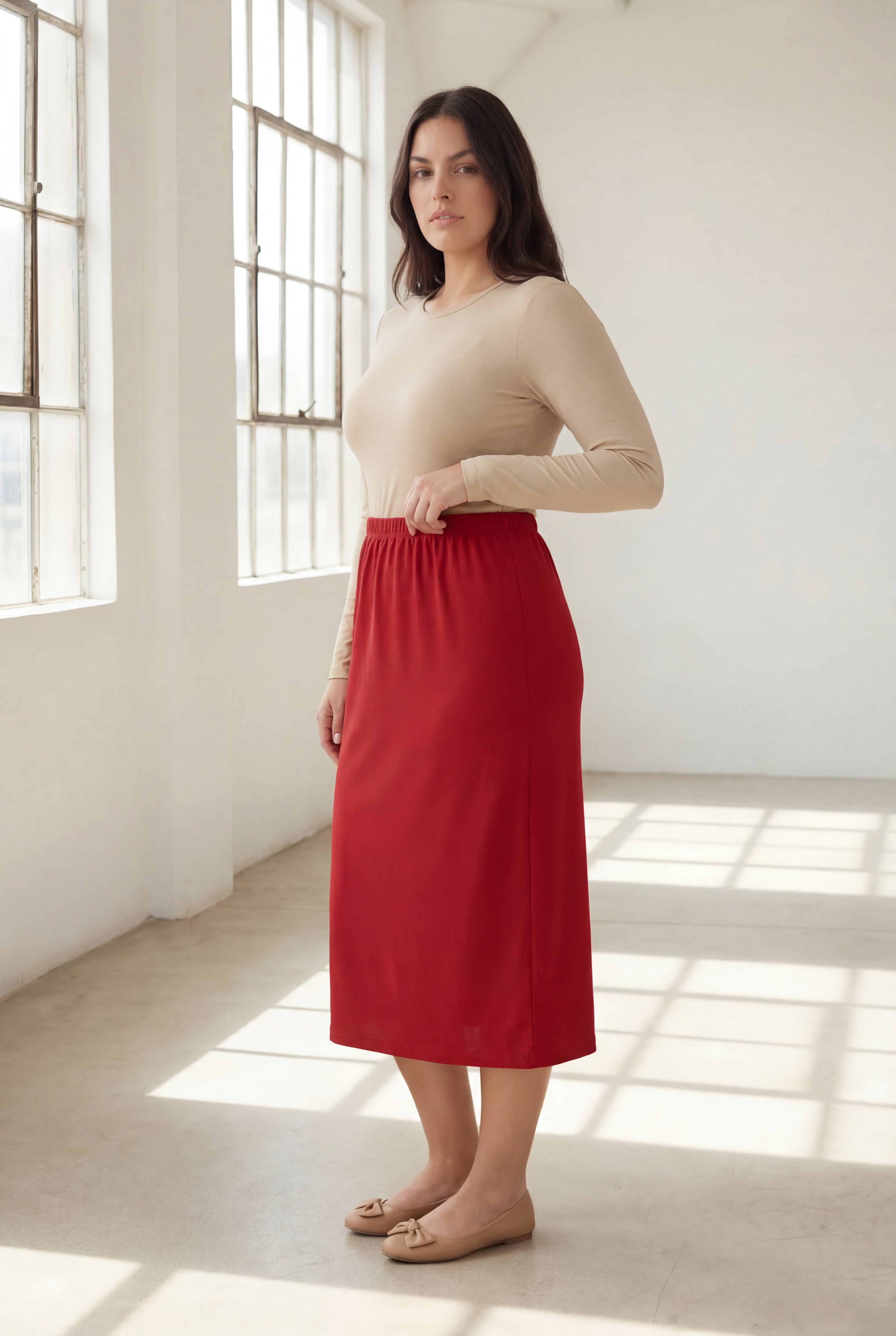 Woman wearing a beige top and red skirt standing in a bright room with large windows.