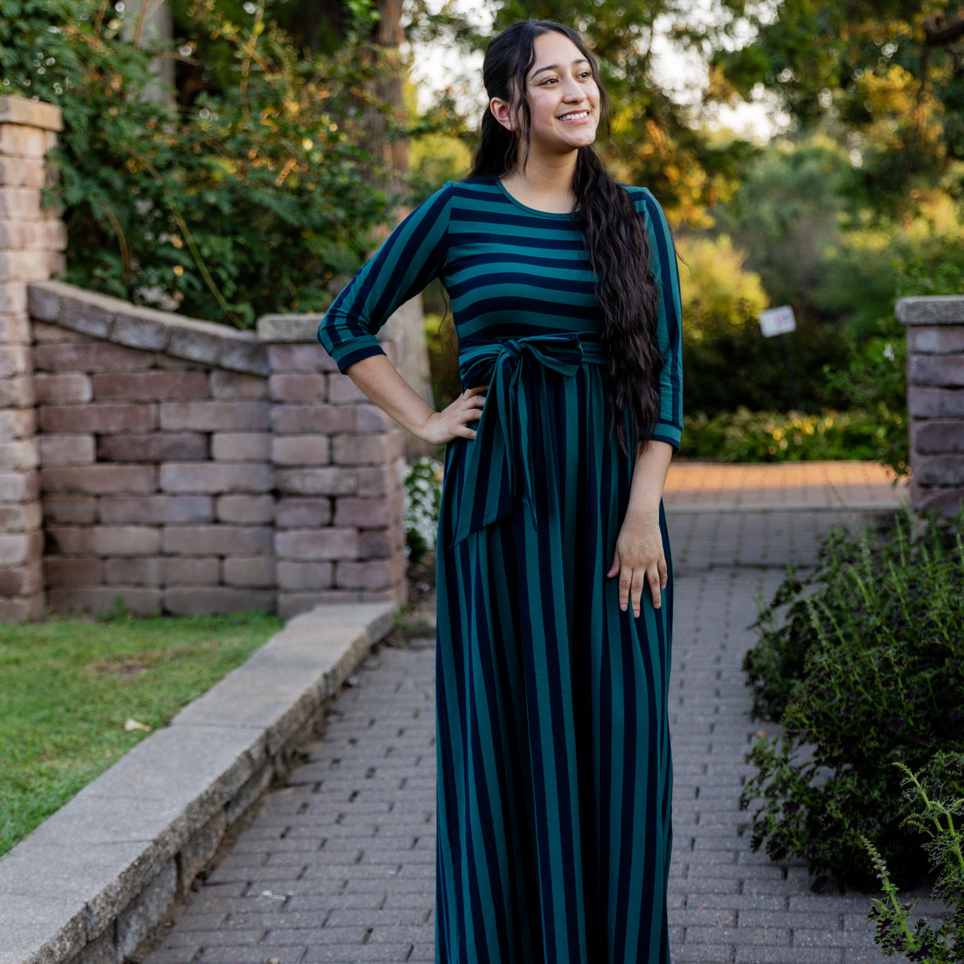 Model wearing the Nuggles® Adelia Dress, a modest striped maxi dress with waist tie and ¾ sleeves.