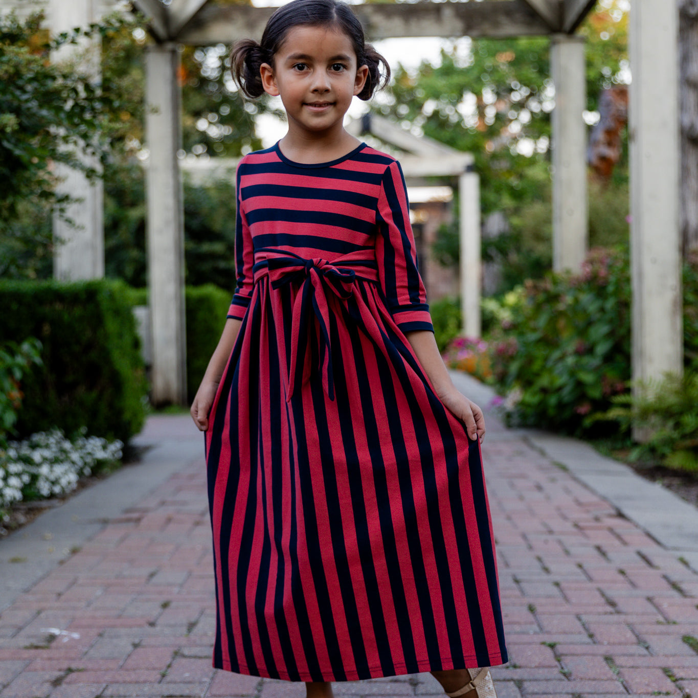 Young girl wearing the Nuggles® Girls Adelia Dress — a modest striped maxi dress with bow tie waist.
