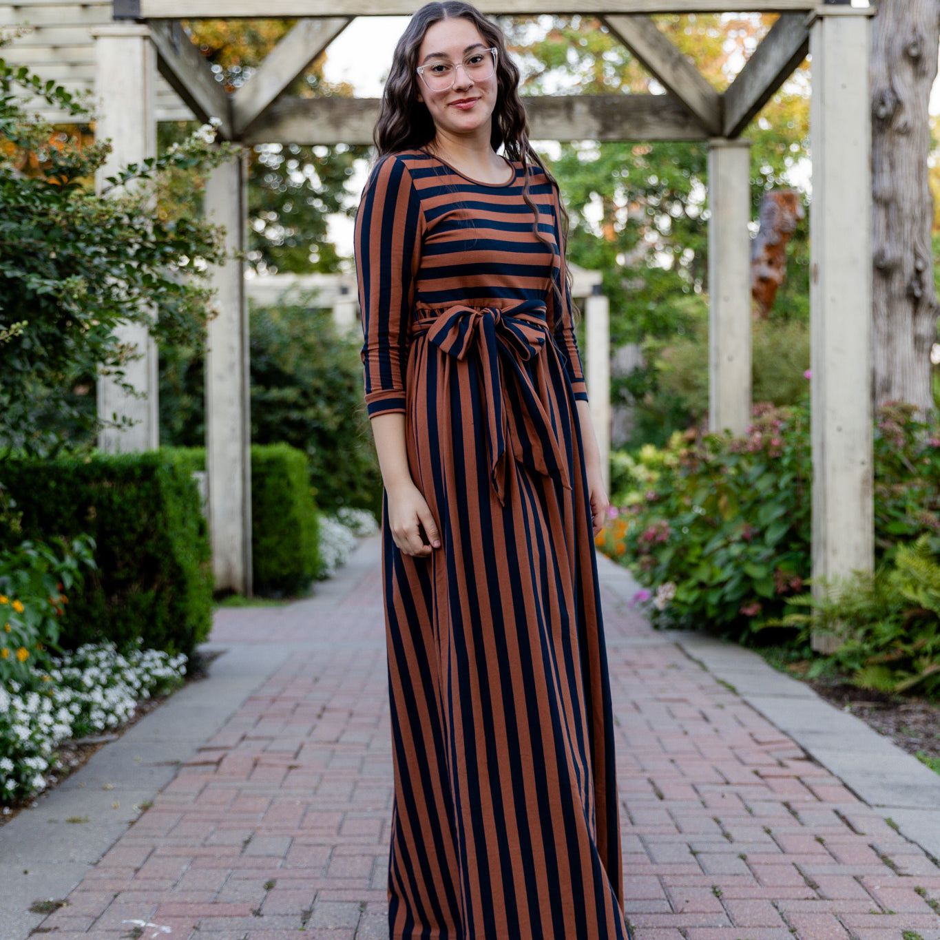 Model wearing the Nuggles® Adelia Dress, a modest striped maxi dress with waist tie and ¾ sleeves.