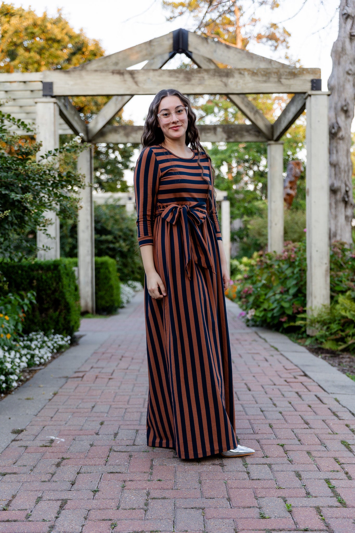 Model wearing the Nuggles® Adelia Dress, a modest striped maxi dress with waist tie and ¾ sleeves.