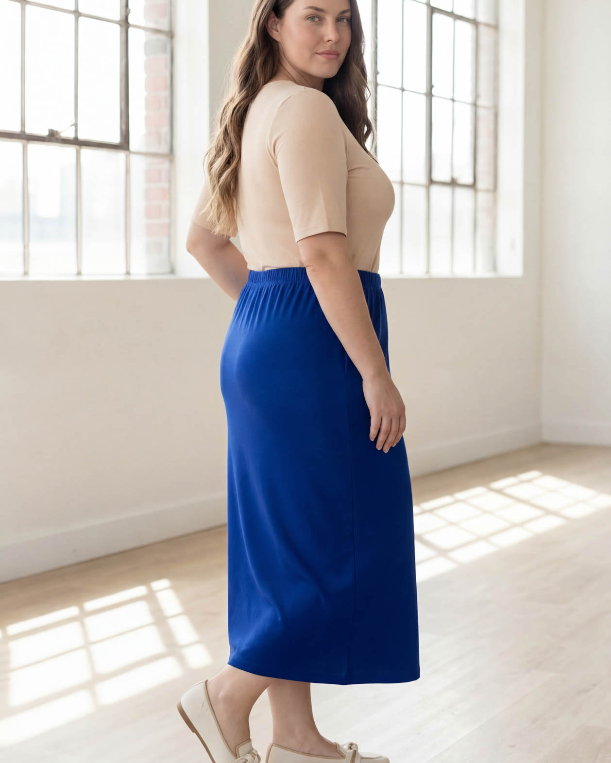 Woman wearing a blue skirt and beige top in a bright room with large windows.