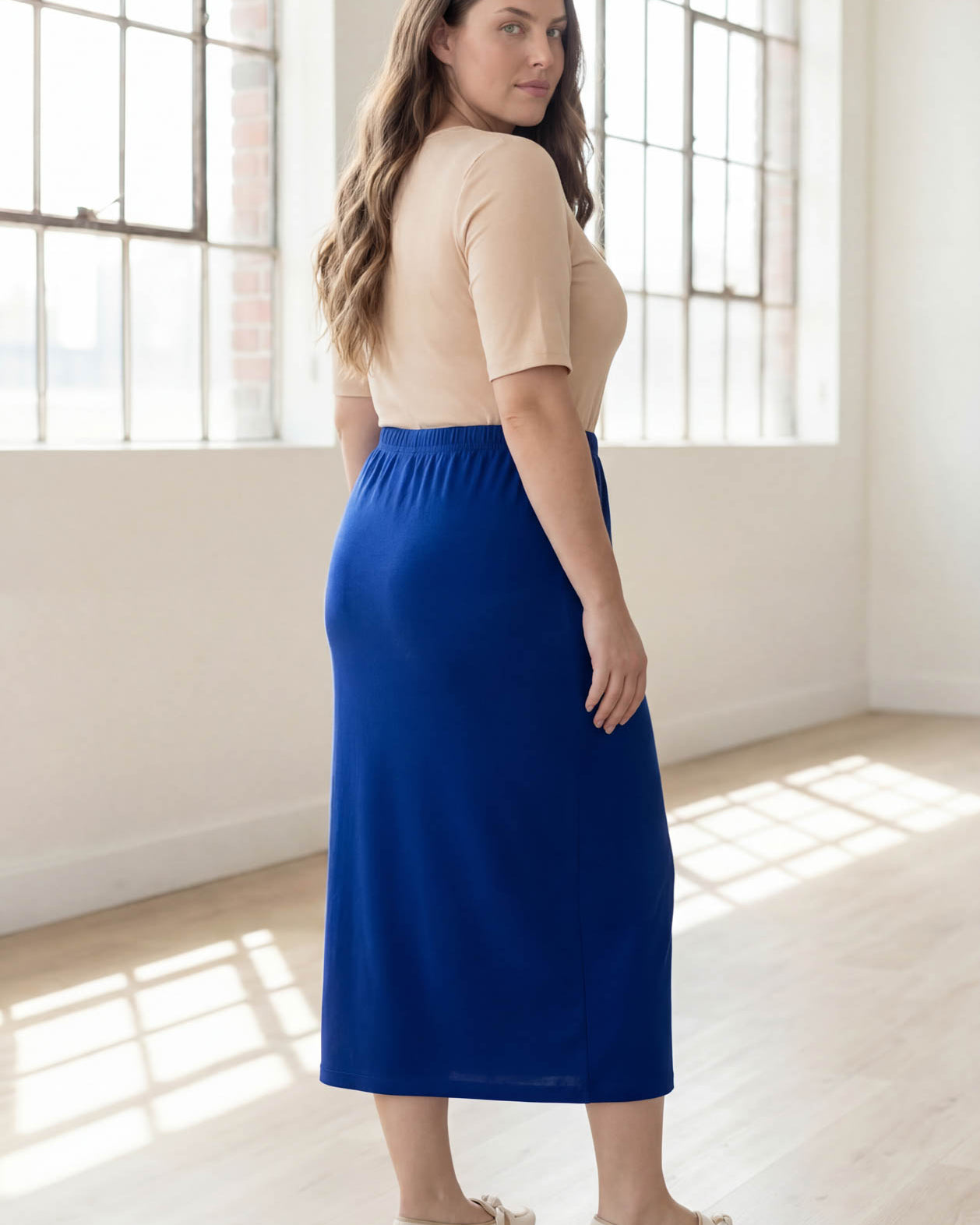 Woman wearing a blue skirt and beige top in a bright room with large windows.