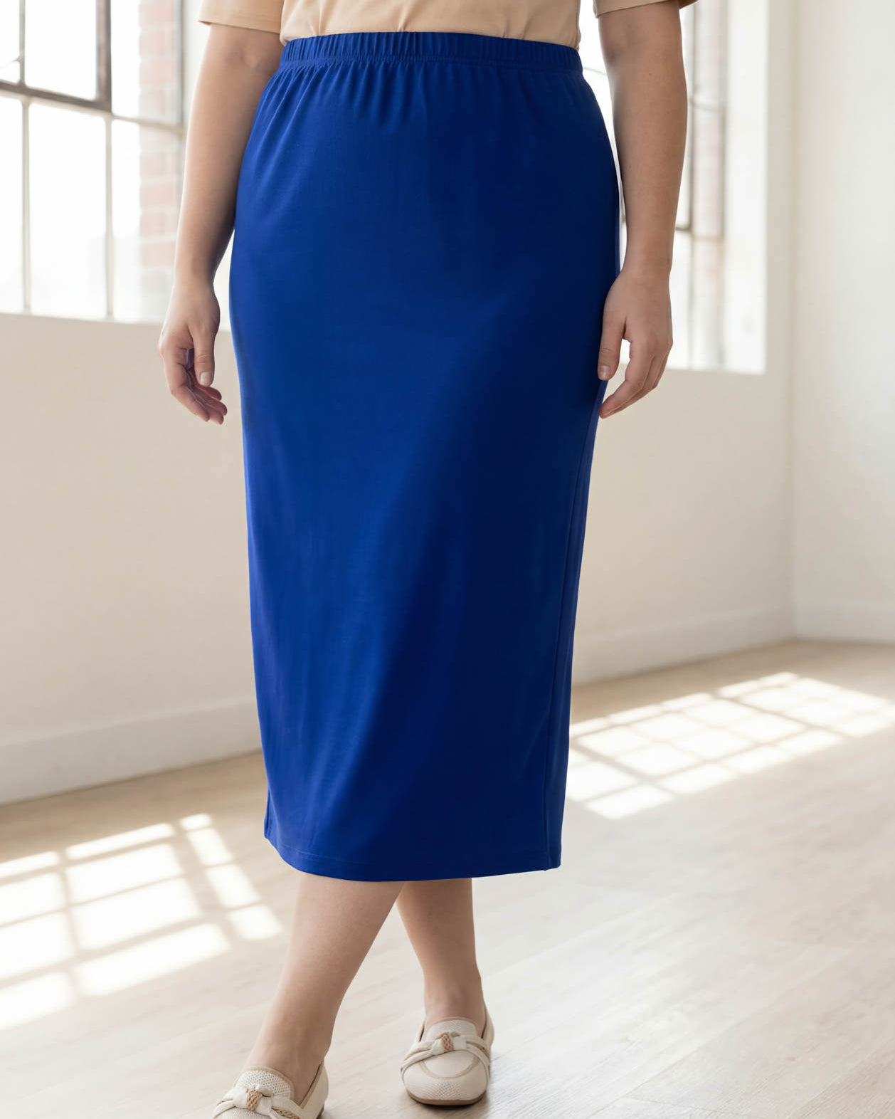 Person wearing a blue skirt and beige top in a bright room with large windows.