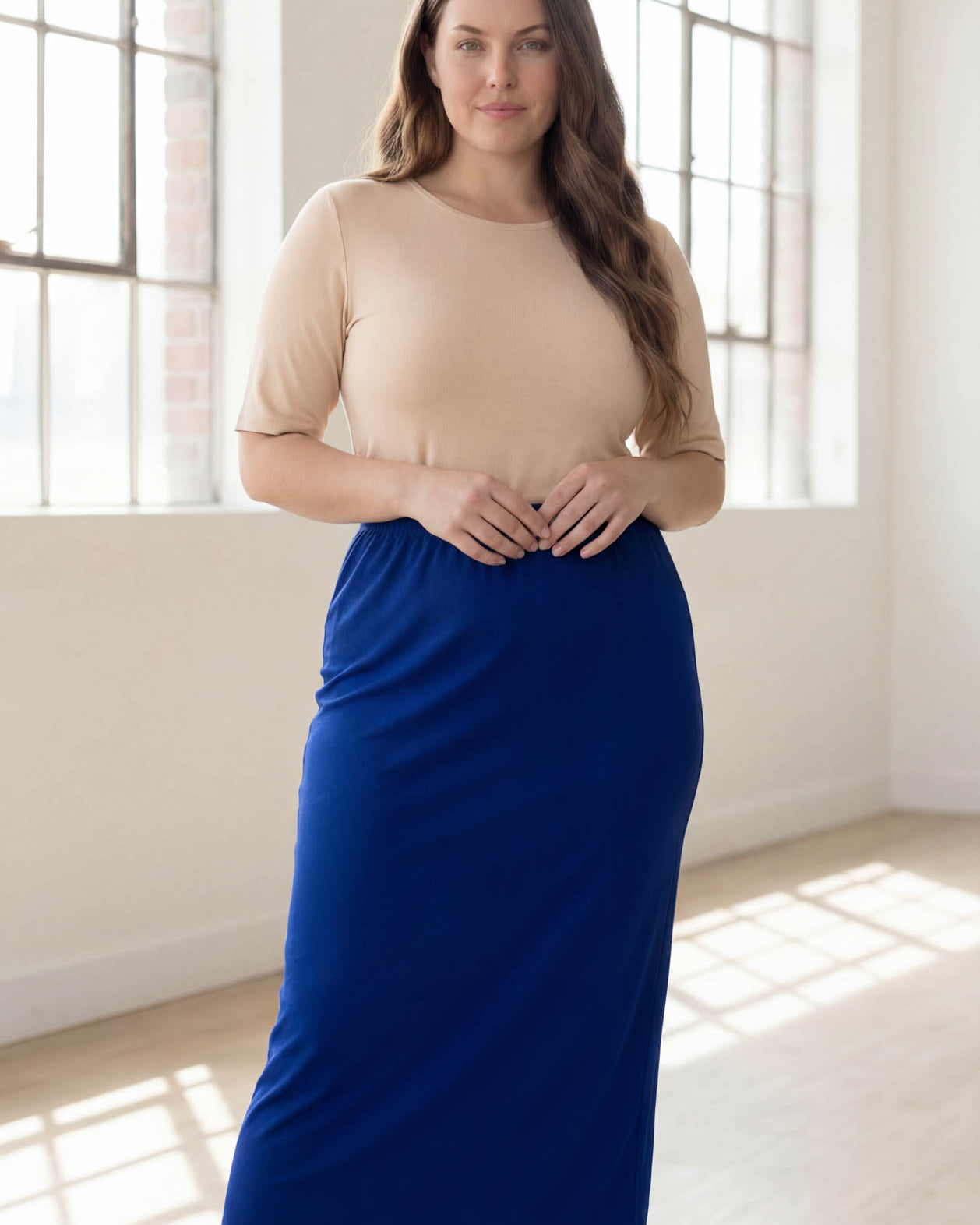 Woman wearing a beige top and blue skirt standing in a bright room with large windows.