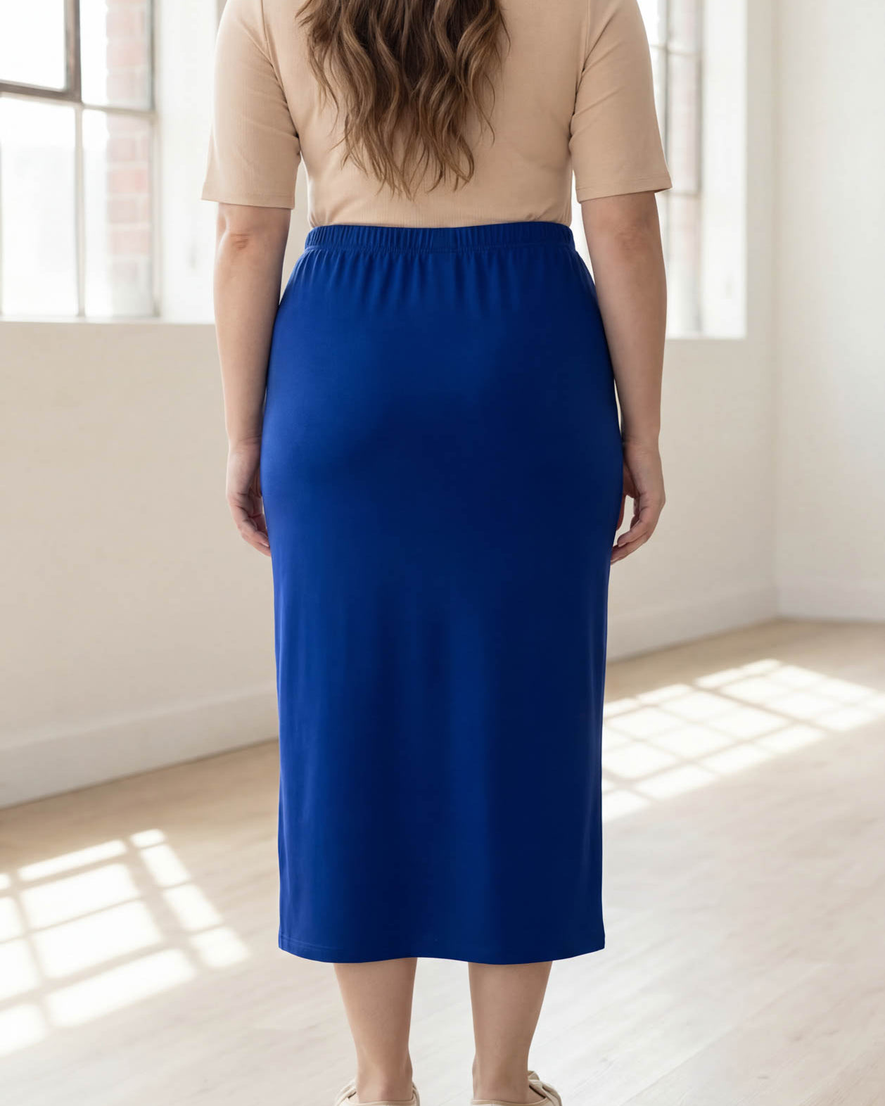 Person wearing a blue skirt and beige top in a bright room with large windows.