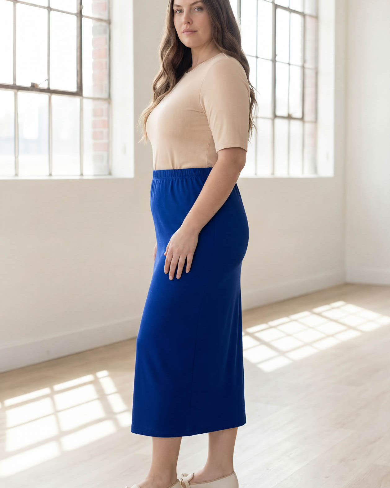 Woman wearing a blue skirt and beige top in a bright room with large windows.