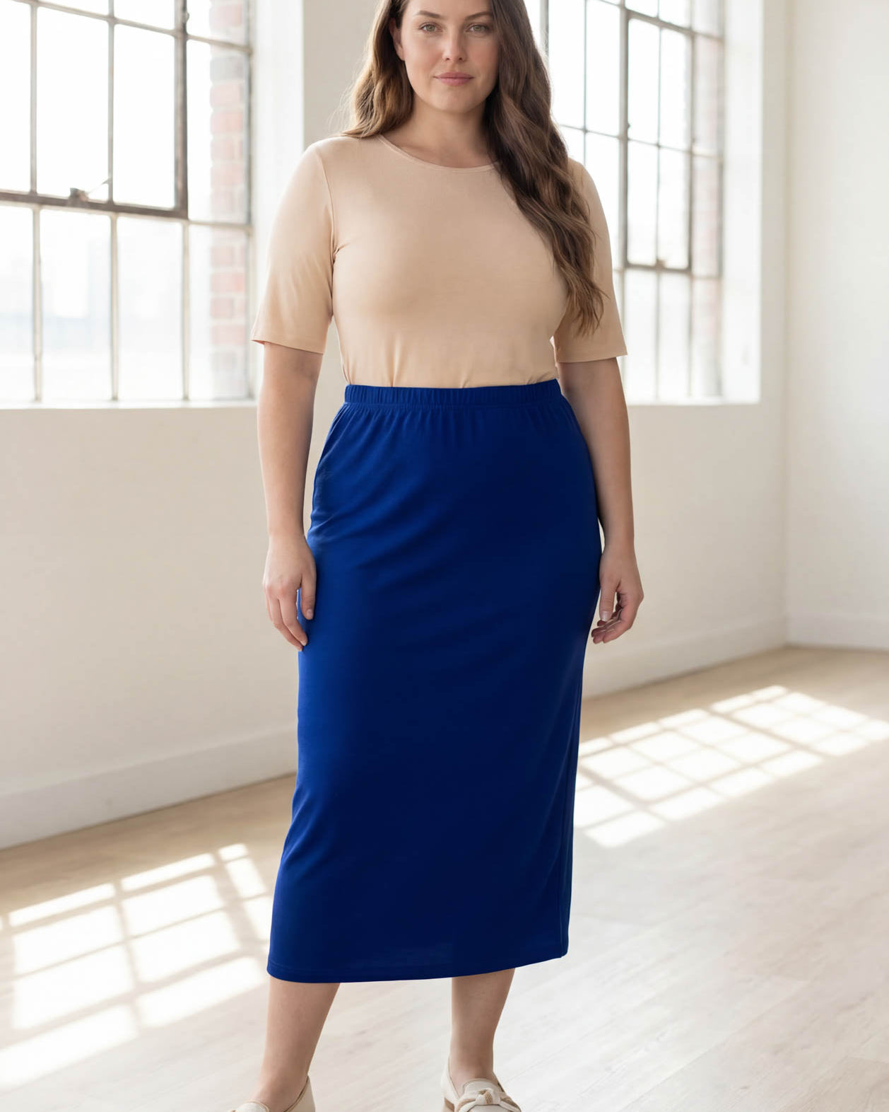 Woman wearing a beige top and blue skirt in a bright room with large windows.