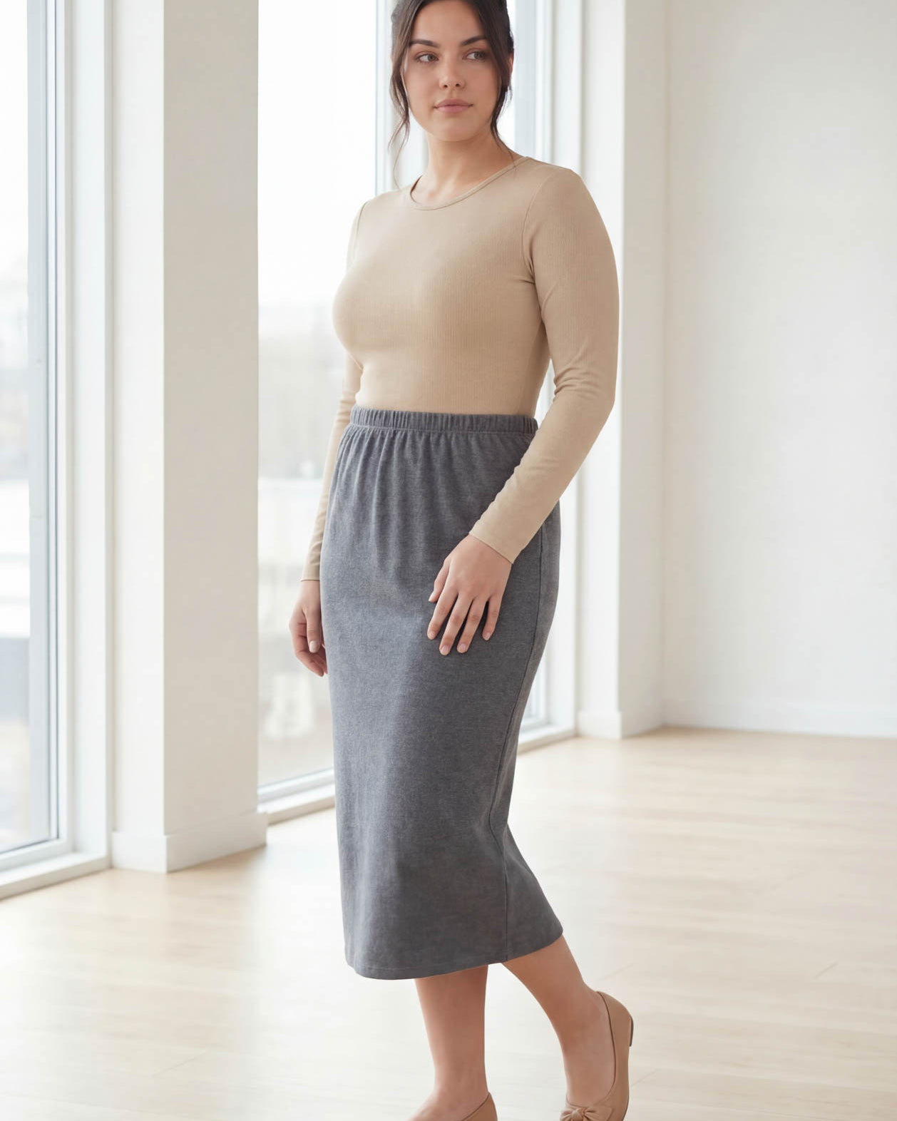 Woman wearing a beige top and gray skirt standing in a bright room with large windows.