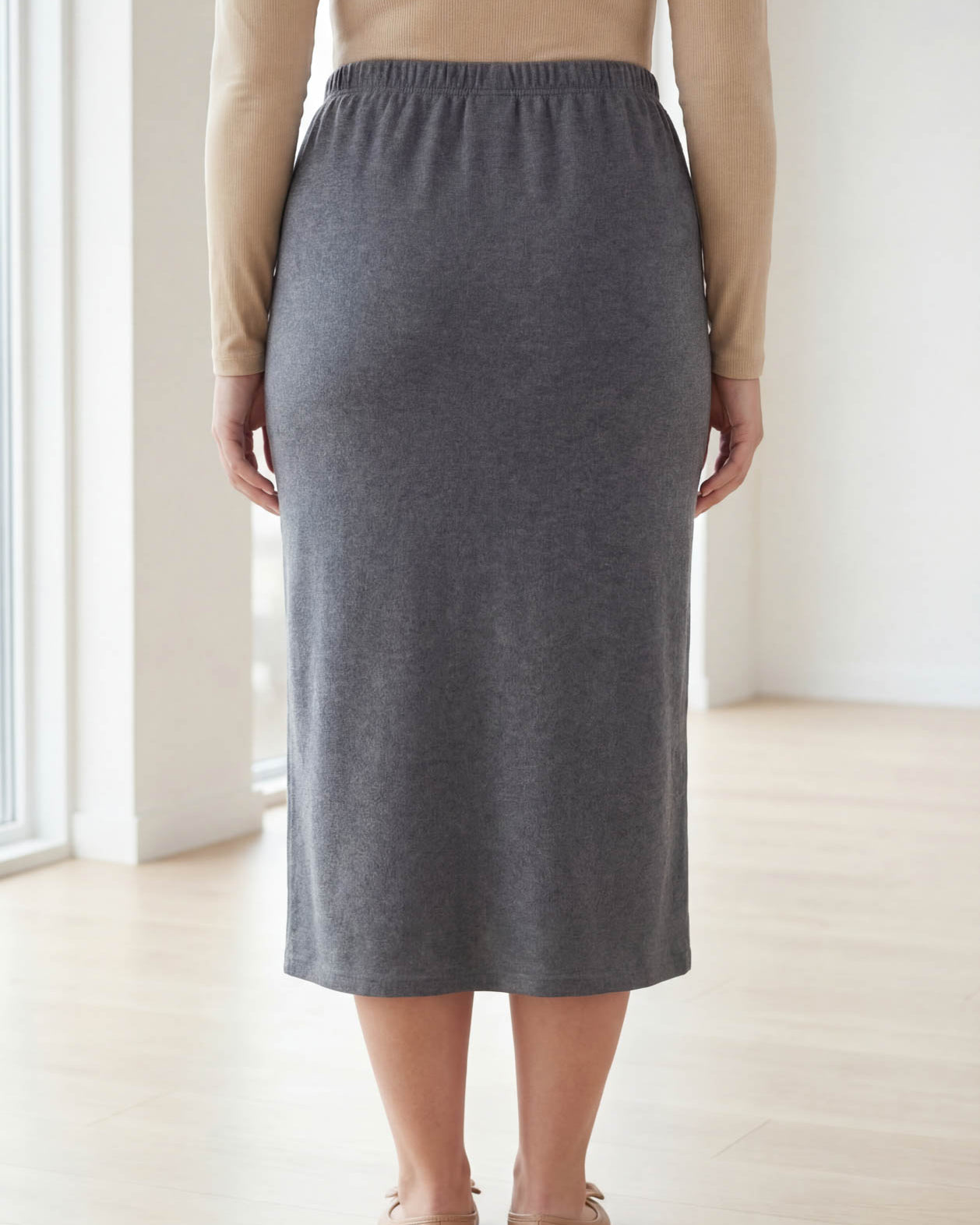 Woman wearing a beige top and gray skirt standing in a bright room with large windows.