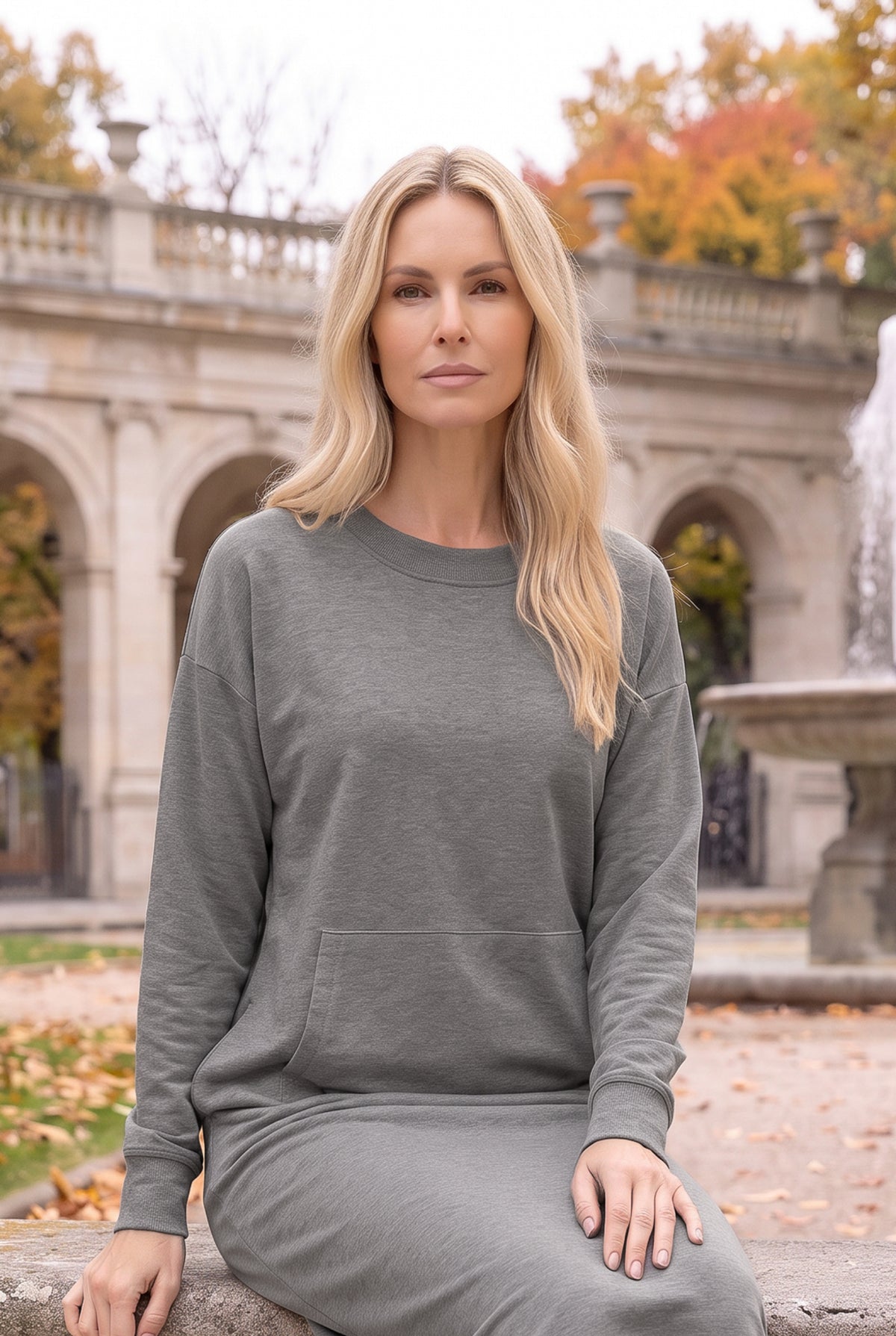 Nuggles® Oakley Hoodie Dress in Heather Gray, seated on a stone ledge with a fountain and fall trees behind her
