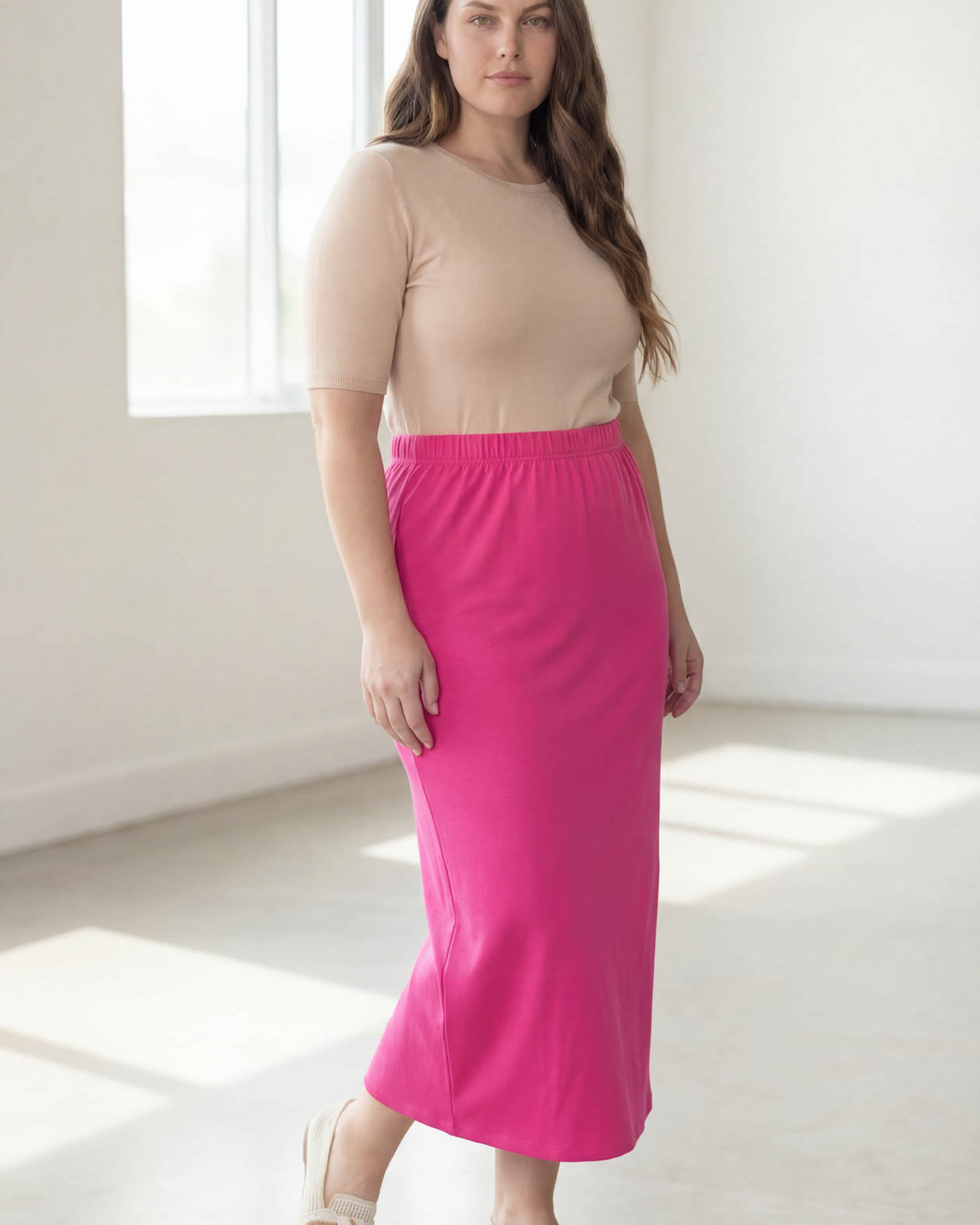 Woman wearing a beige top and pink skirt in a bright room.