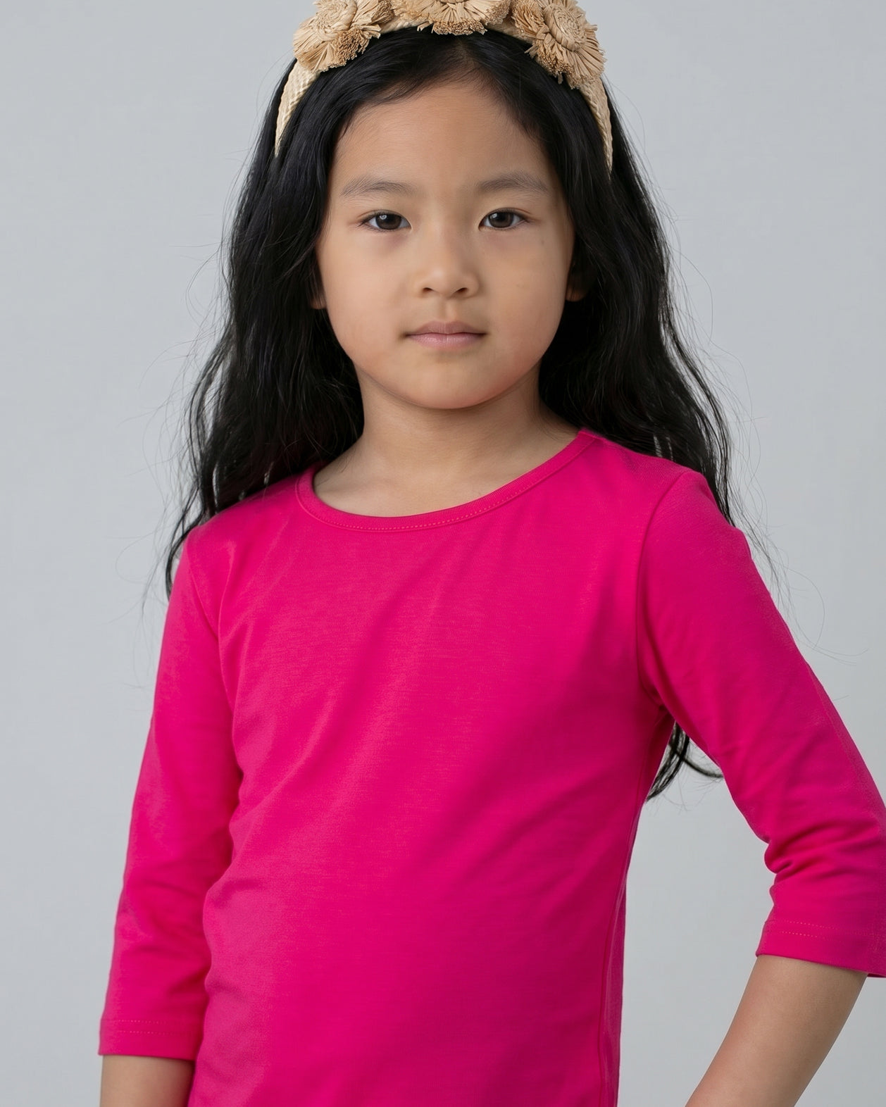 Young girl wearing a bright pink long-sleeve shirt and beige headband with flowers on a gray background