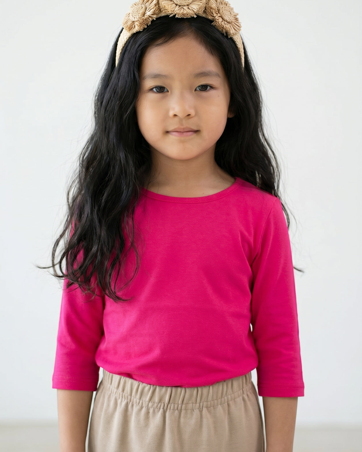 Young girl wearing a bright pink long-sleeve shirt and beige headband with flowers on a gray background