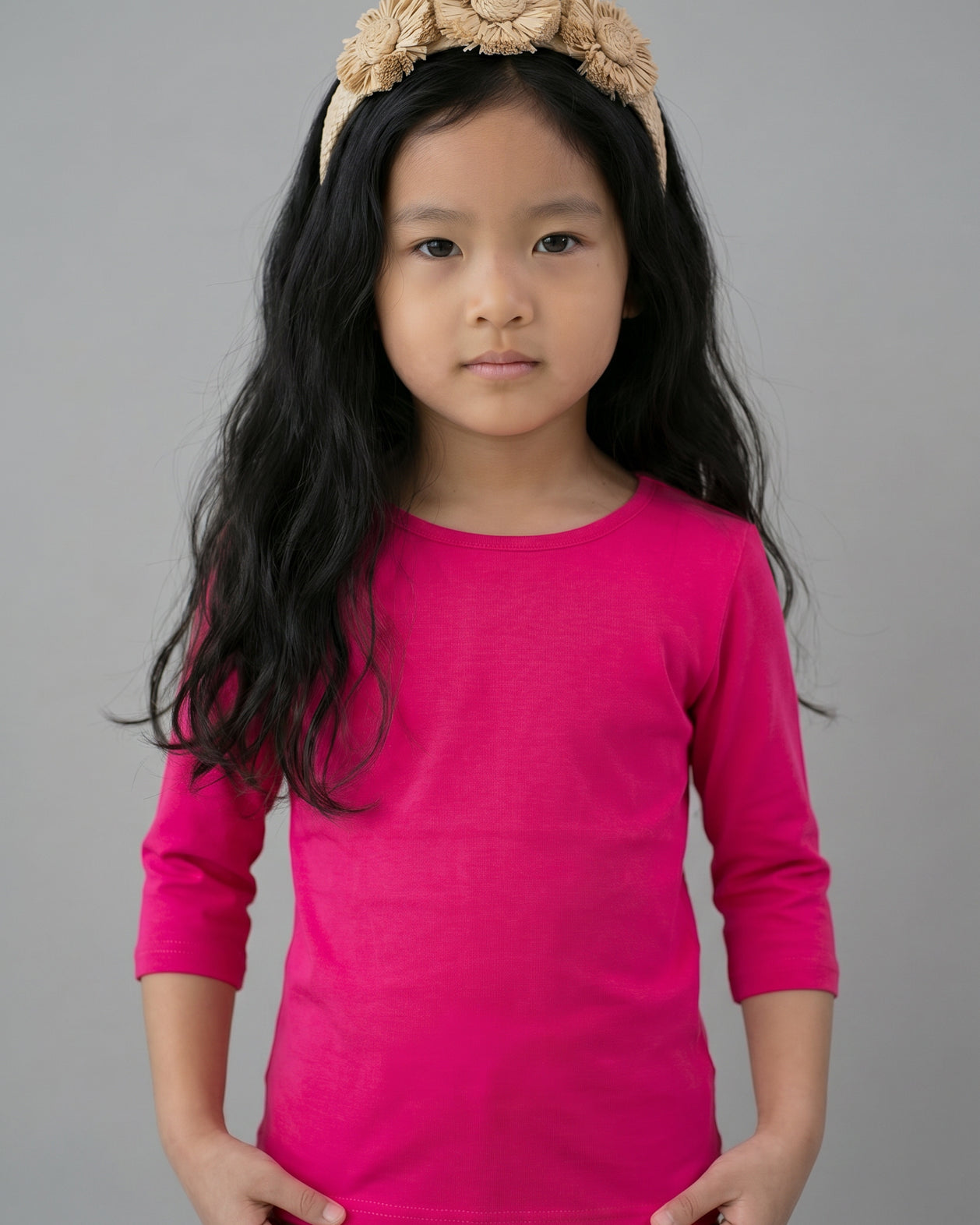 Young girl wearing a bright pink long-sleeve shirt and beige headband with flowers on a gray background
