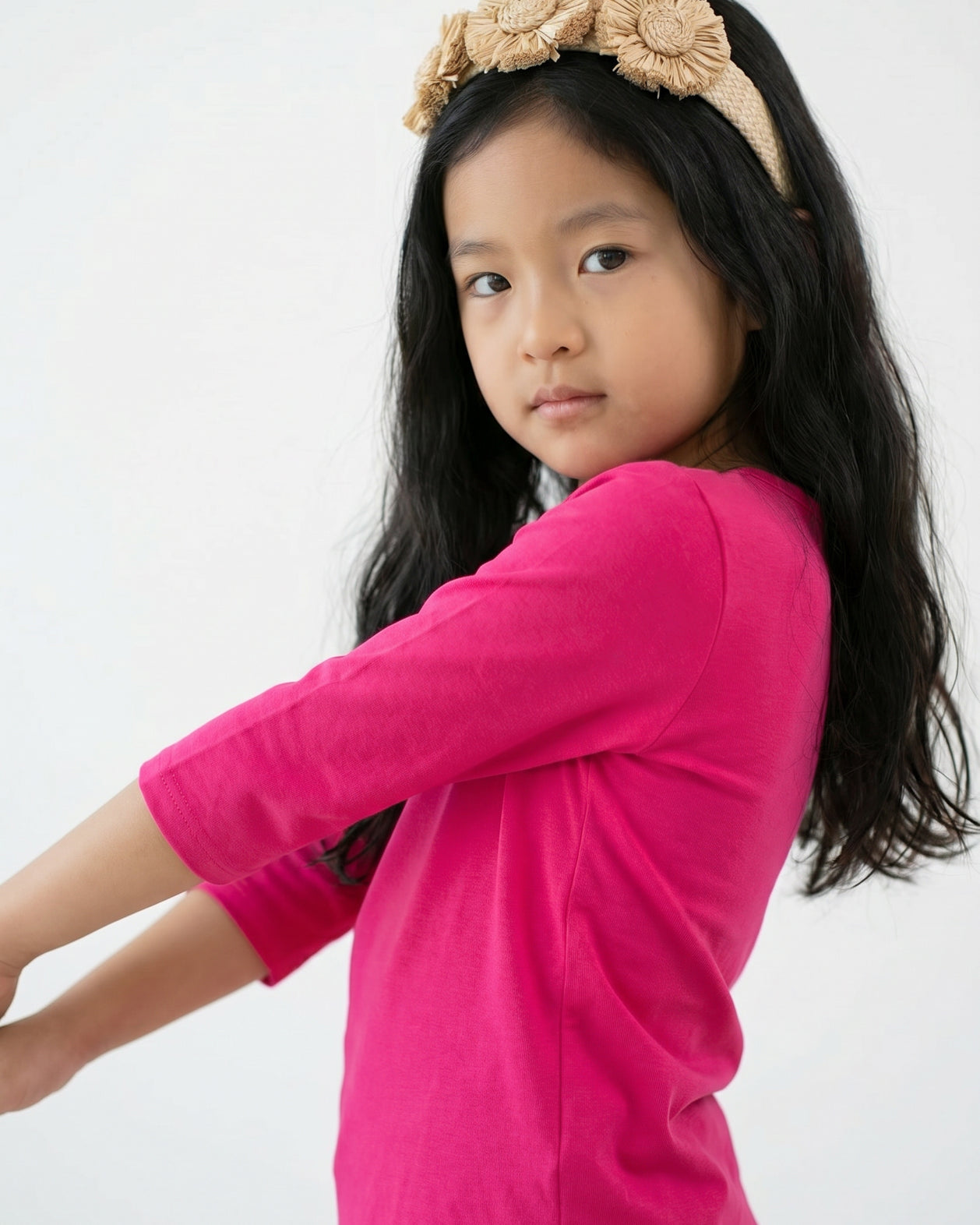 Young girl wearing a bright pink long-sleeve shirt and beige headband with flowers on a gray background