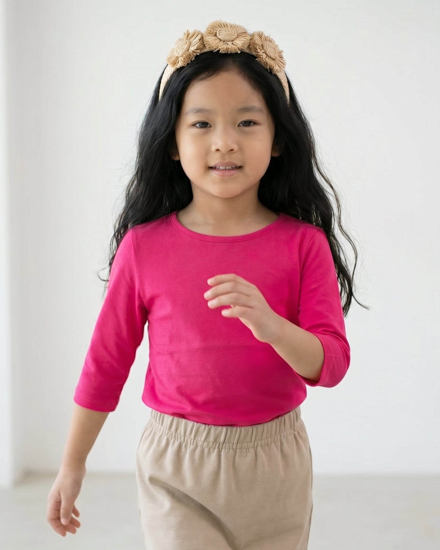 Young girl wearing a bright pink long-sleeve shirt and beige headband with flowers on a gray background