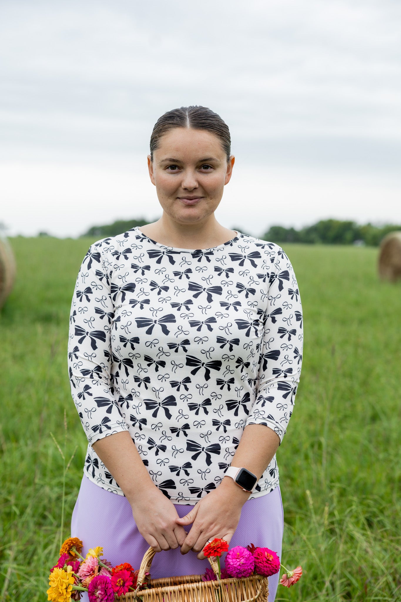 Woman standing in a field holding a basket of flowers, wearing a patterned shirt.