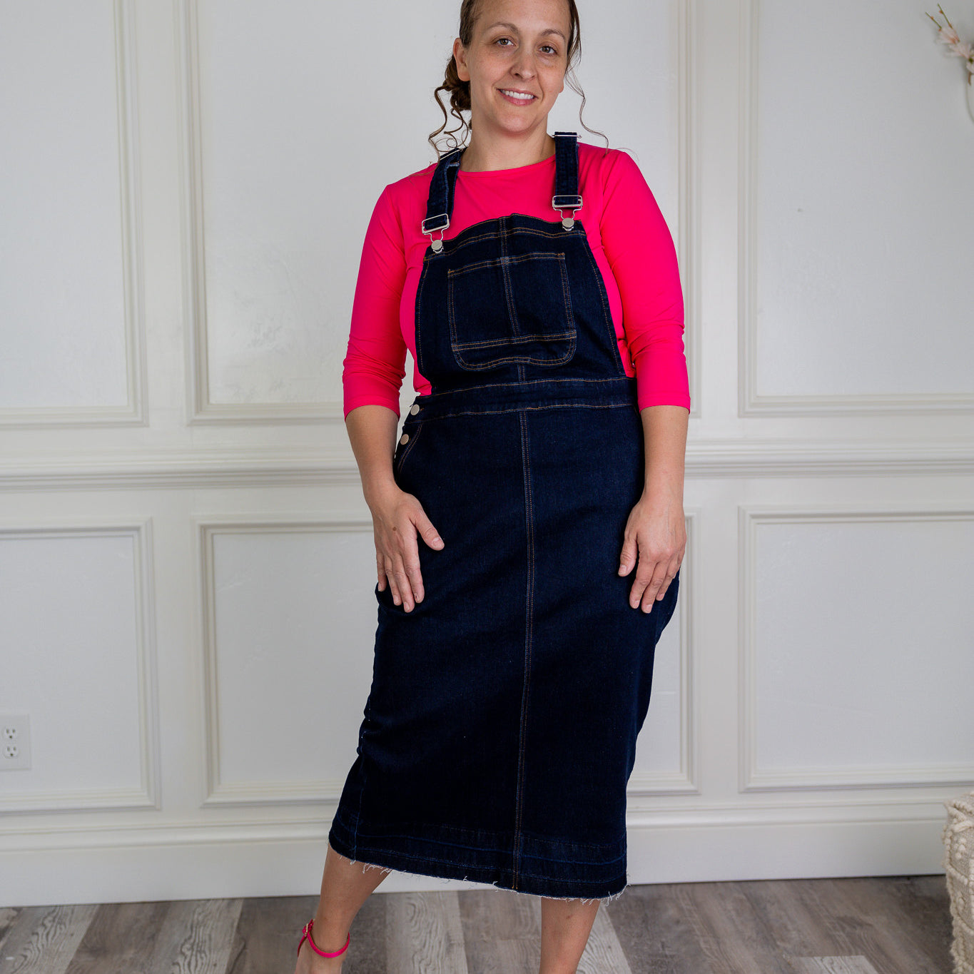 Model wearing a 'NUGGLES® modest dark wash denim overall dress with front pocket and raw hem.