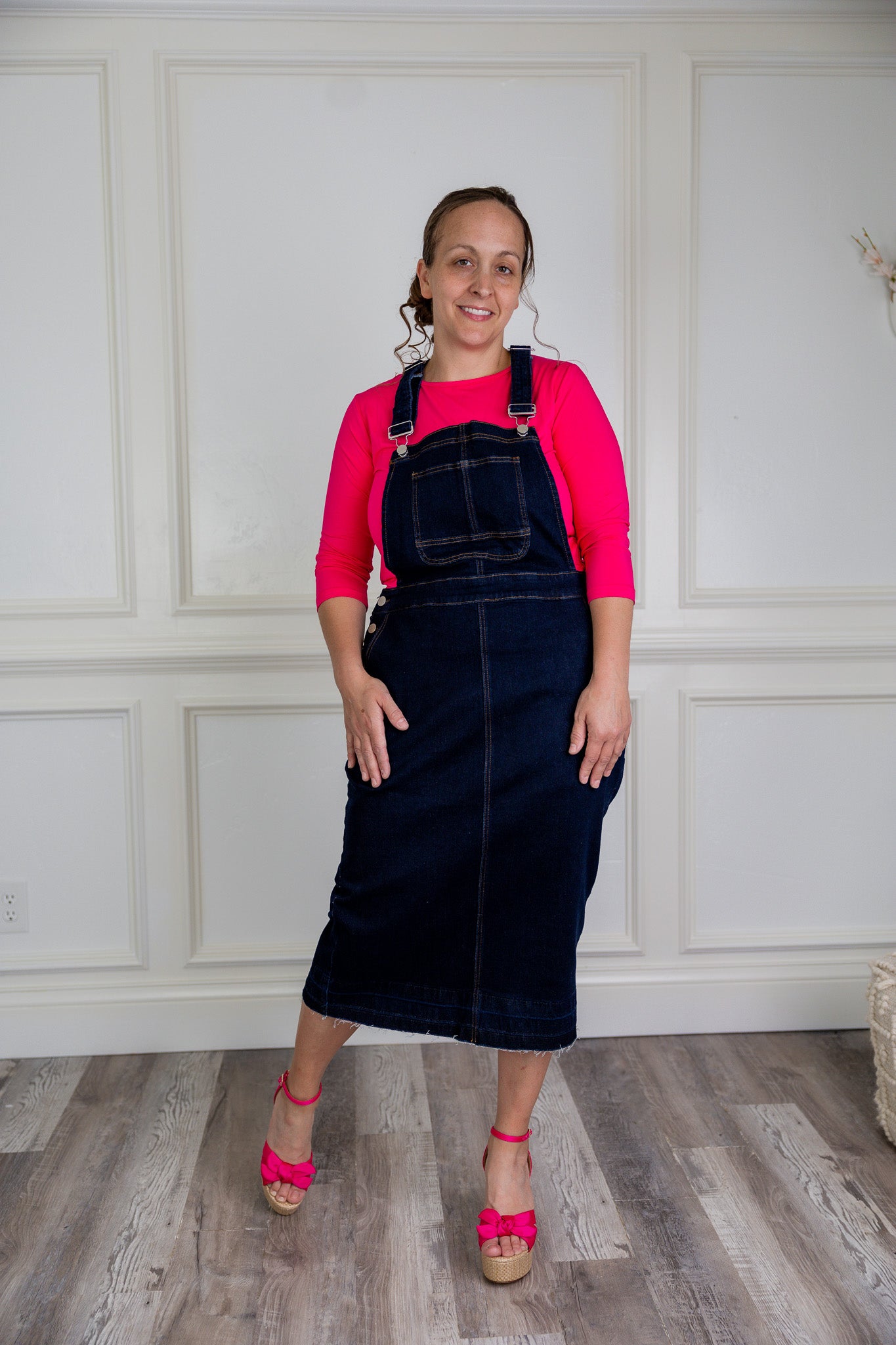 Model wearing a 'NUGGLES® modest dark wash denim overall dress with front pocket and raw hem.