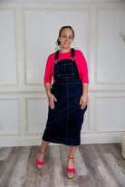 Model wearing a 'NUGGLES® modest dark wash denim overall dress with front pocket and raw hem.