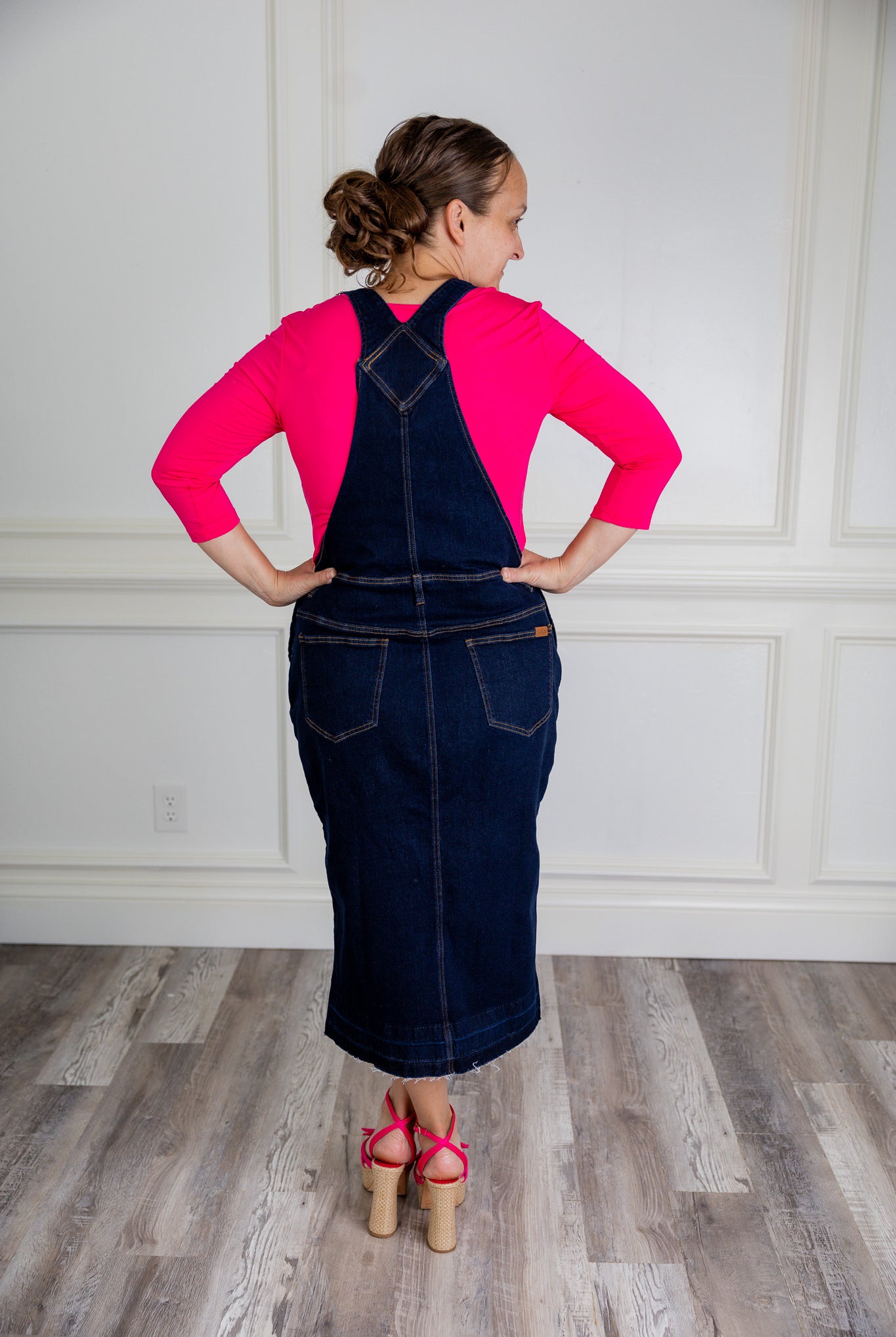 Model wearing a 'NUGGLES® modest dark wash denim overall dress with front pocket and raw hem.