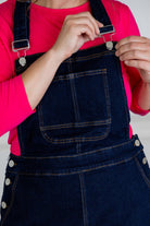 Model wearing a 'NUGGLES® modest dark wash denim overall dress with front pocket and raw hem.
