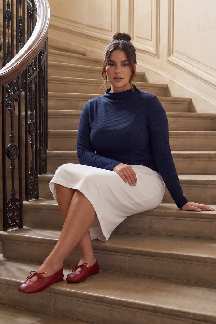 A woman sits on the steps of an elegant indoor staircase, wearing a navy blue mock-neck sweater, a white knee-length skirt, and red Mary Jane shoes.