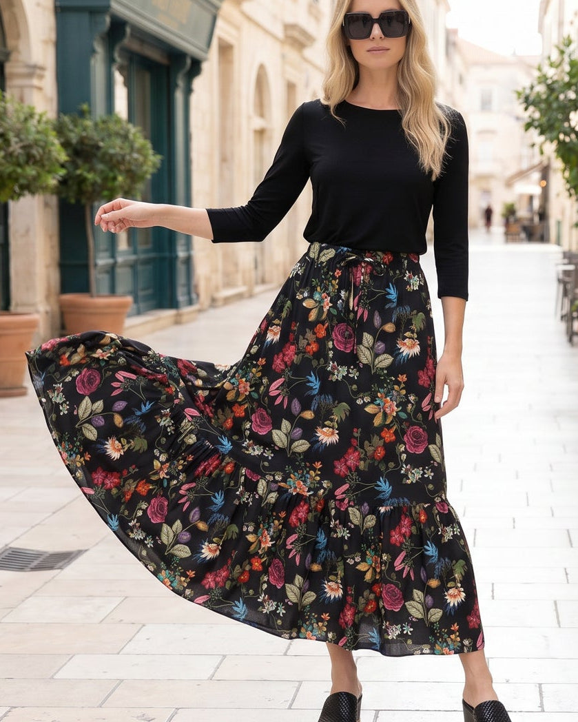Woman in a black top and floral skirt standing on a street with potted plants and buildings.