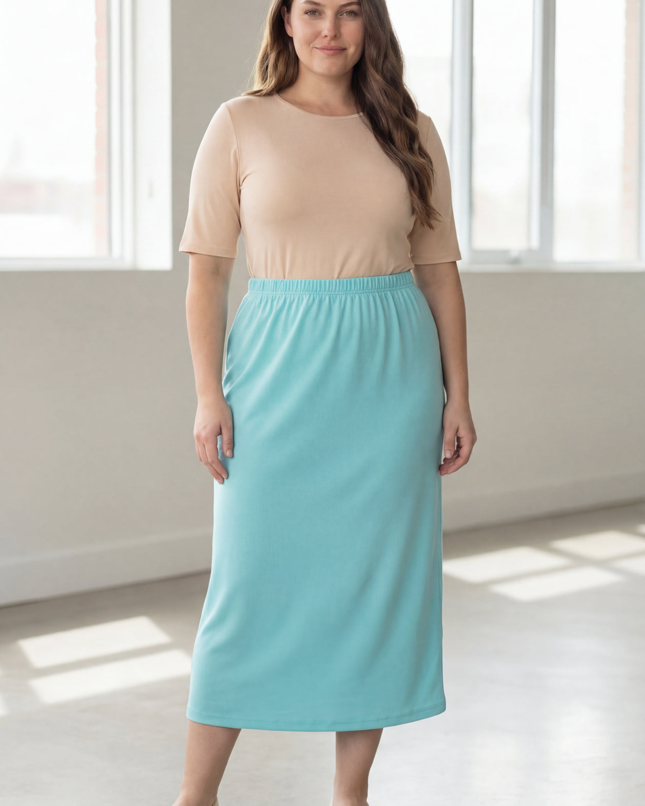 Woman wearing a beige shirt & light blue maxi length pencil skirt standing in a bright room with large windows.
