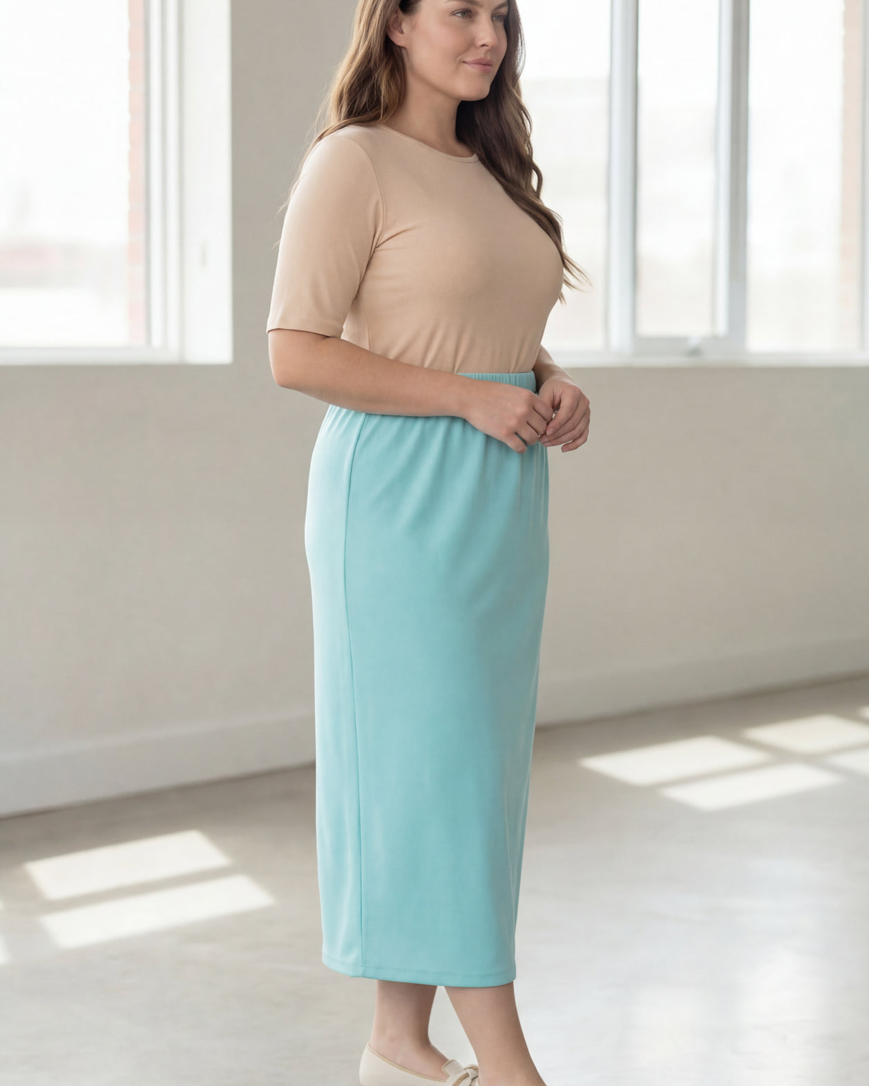 Woman wearing a beige shirt & light blue maxi length pencil skirt standing in a bright room with large windows.