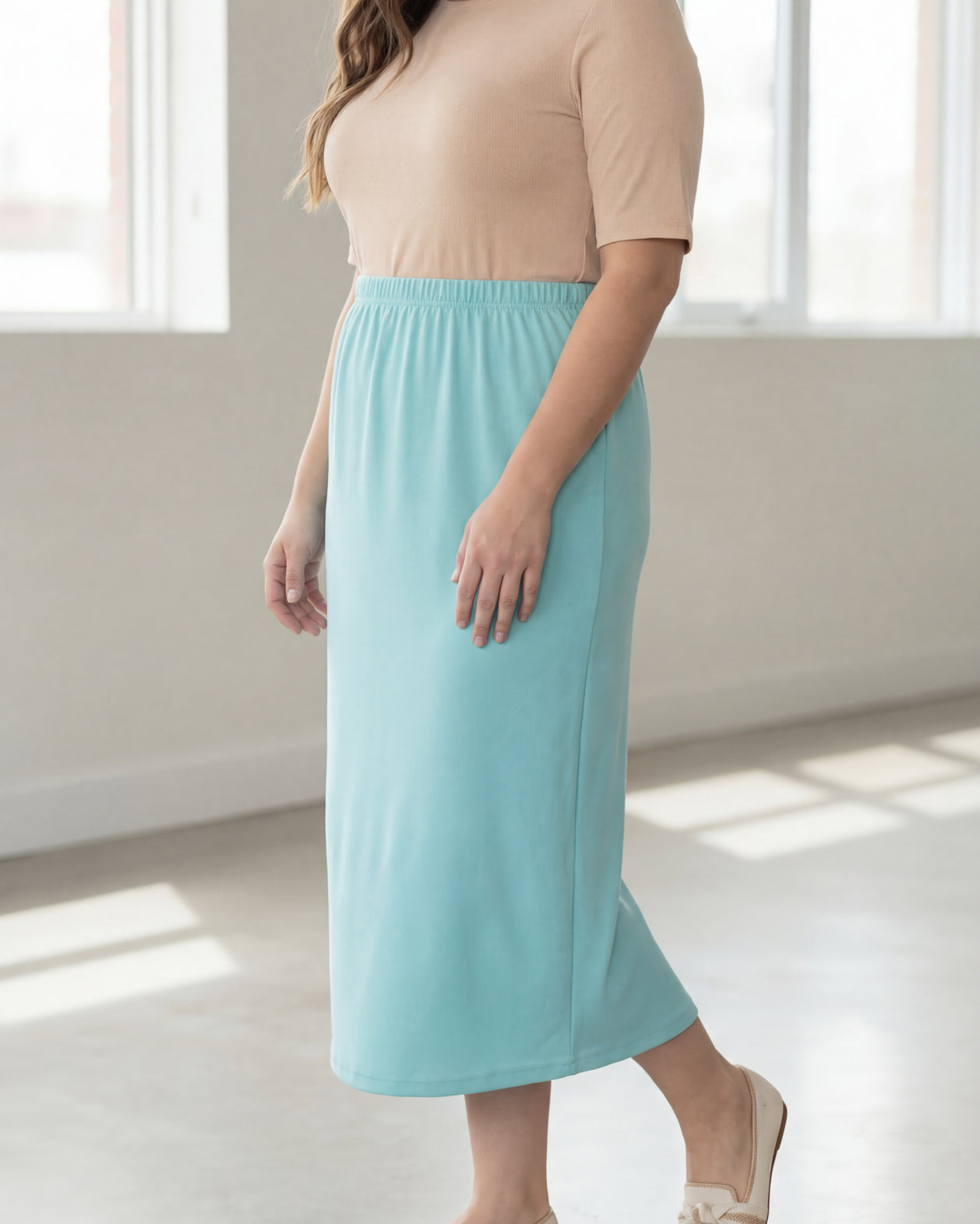 Woman wearing a beige shirt & light blue maxi length pencil skirt standing in a bright room with large windows.