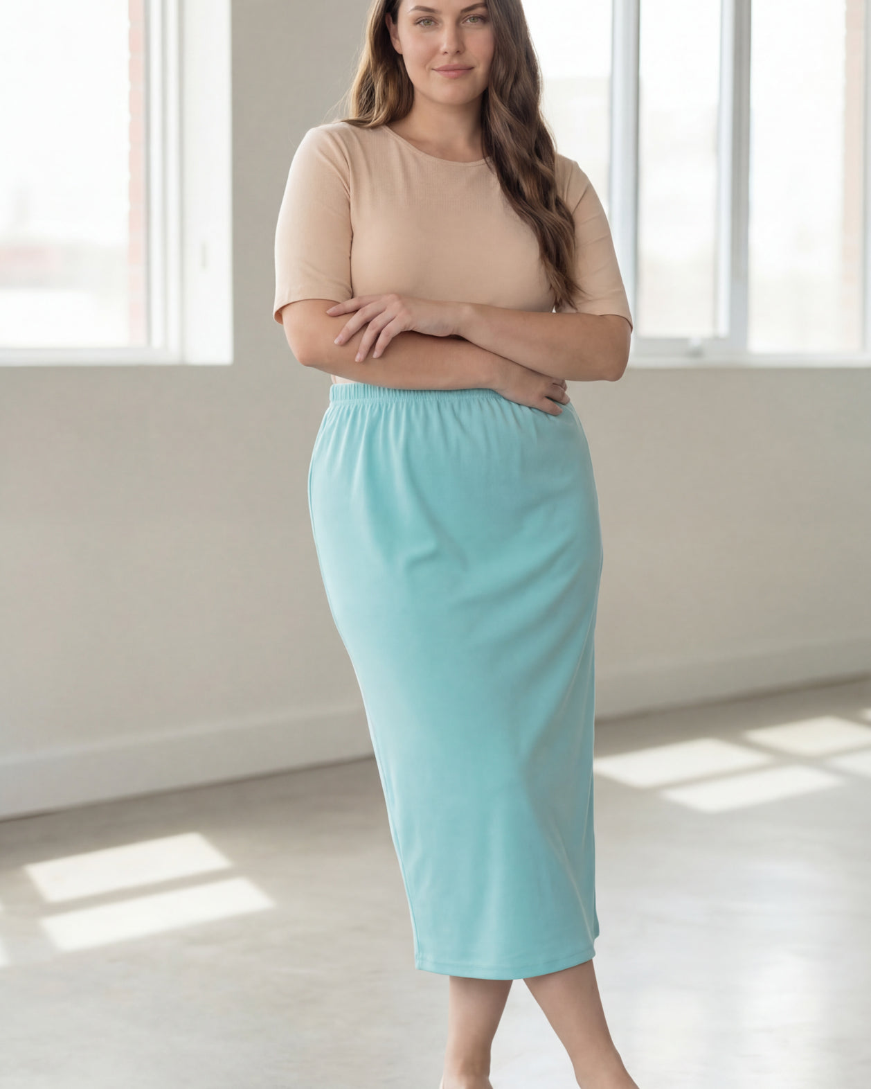 Woman wearing a beige shirt & light blue maxi length pencil skirt standing in a bright room with large windows.