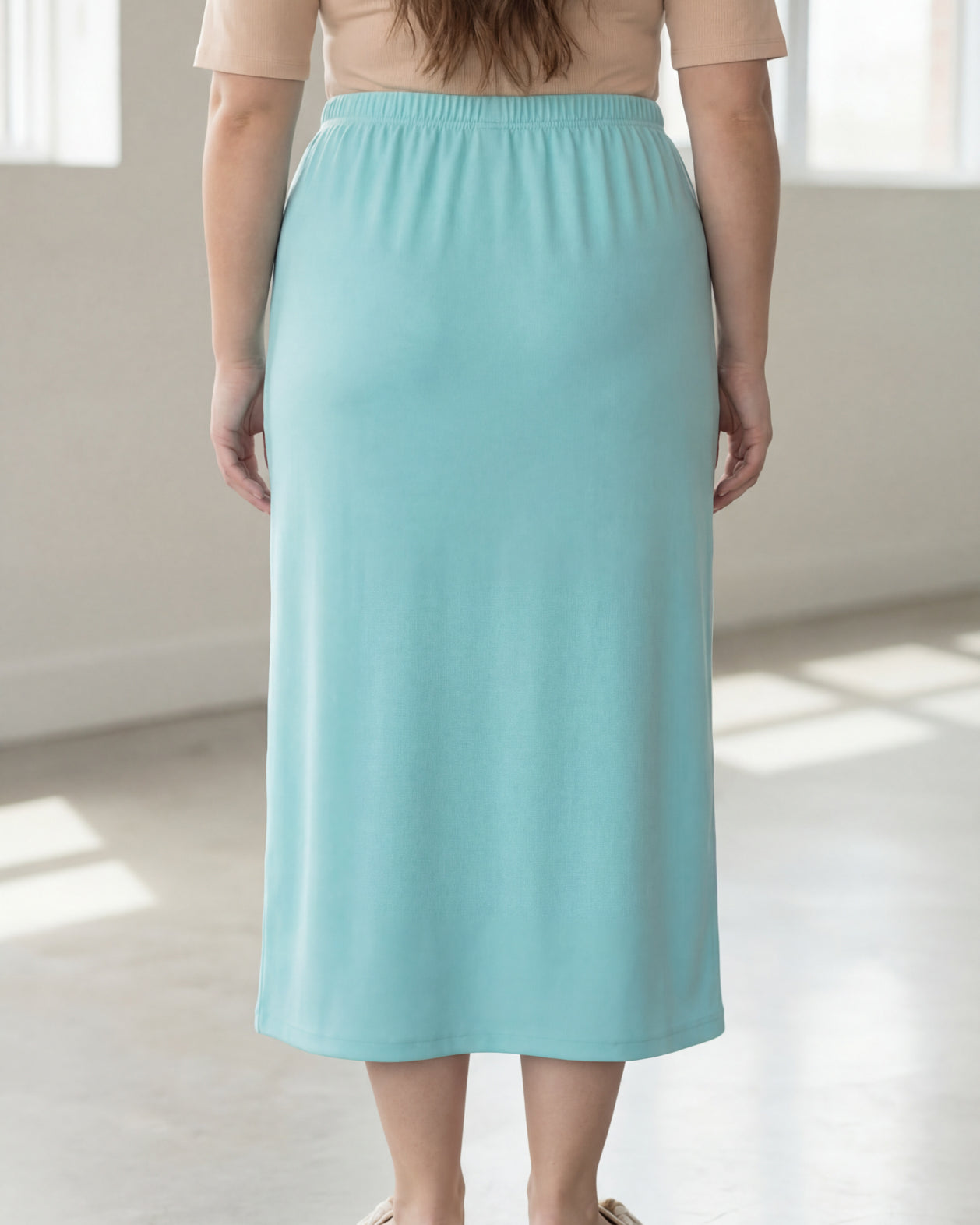 Woman wearing a beige shirt & light blue maxi length pencil skirt standing in a bright room with large windows.