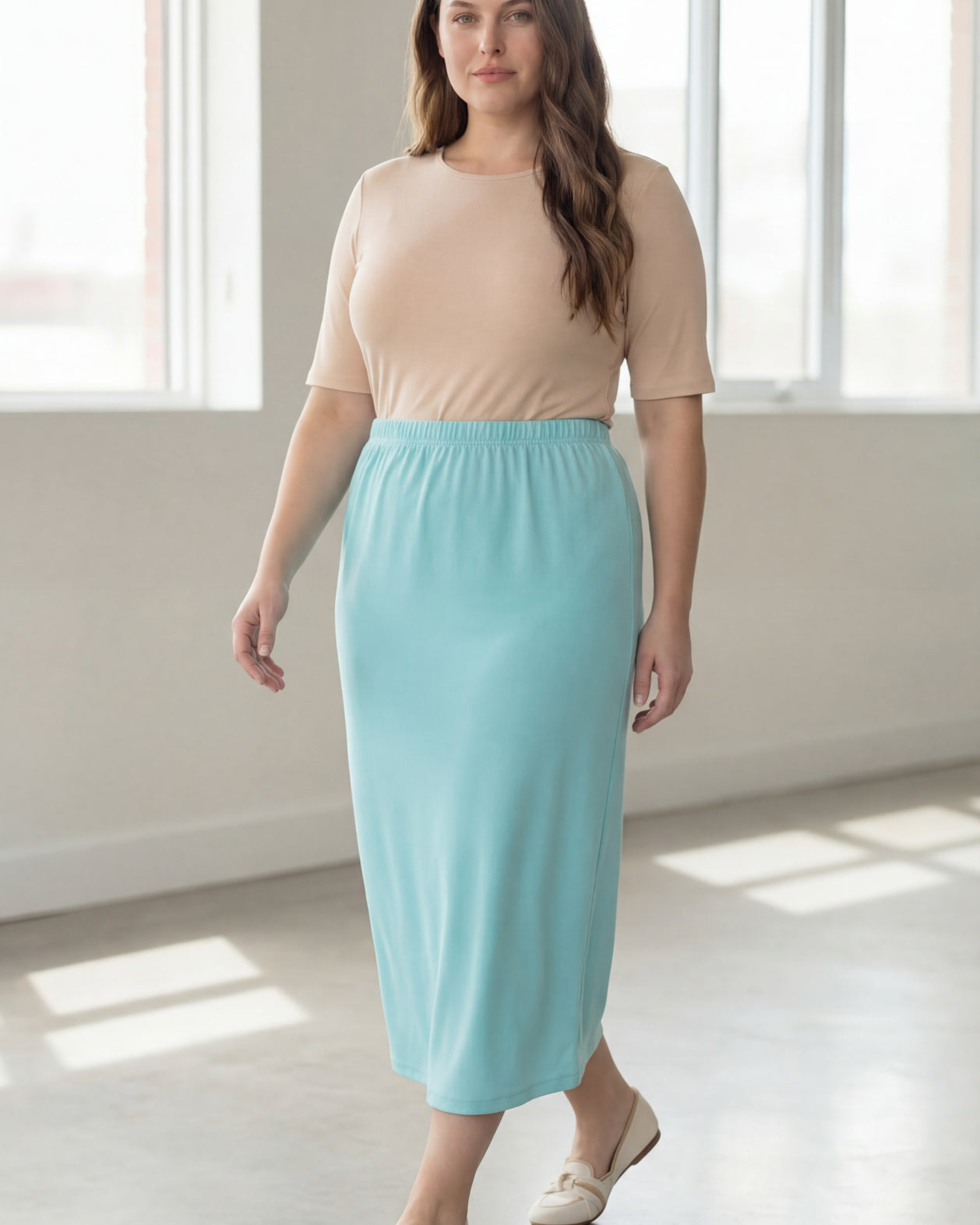 Woman wearing a beige shirt & light blue maxi length pencil skirt standing in a bright room with large windows.