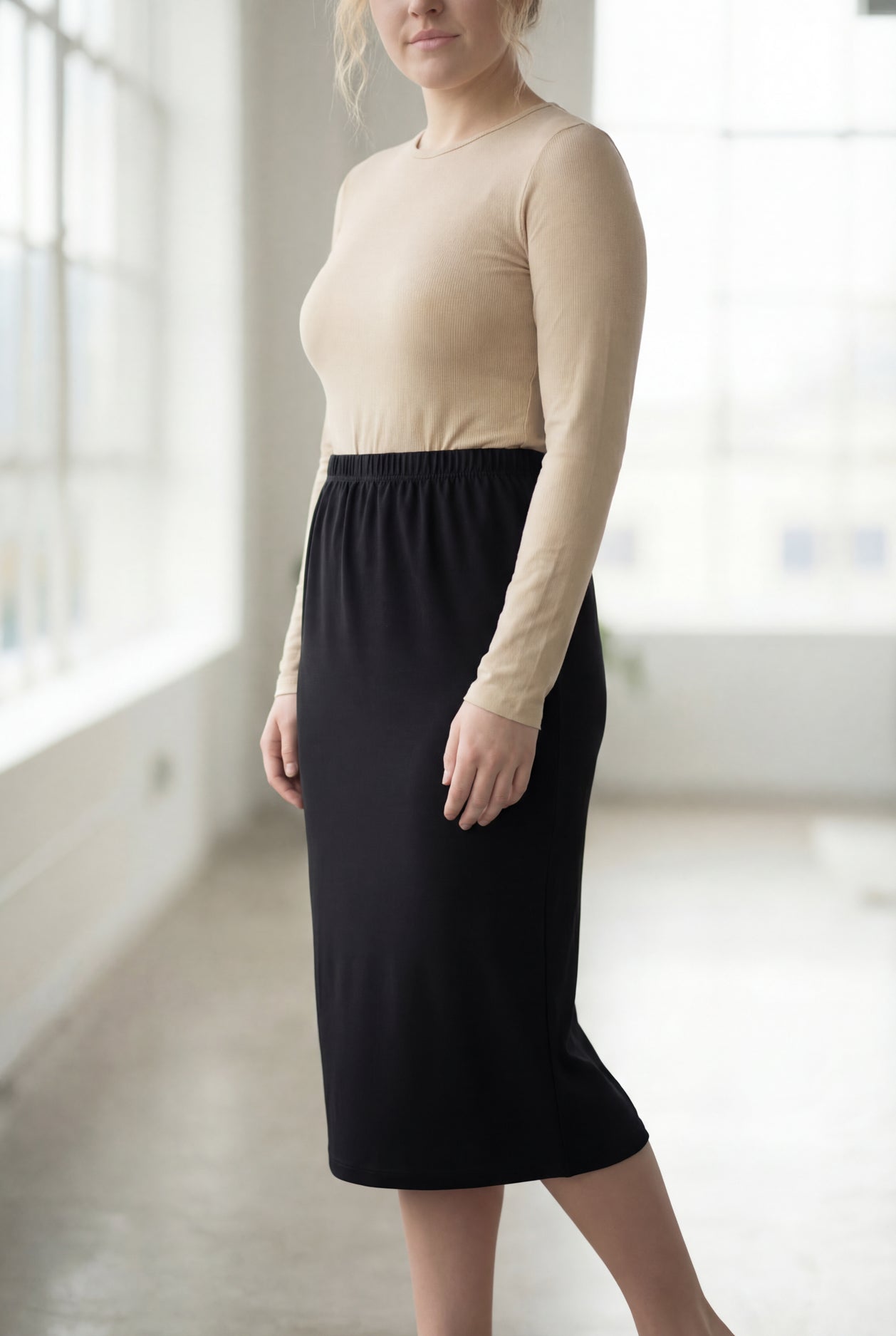Women wearing a solid black classic 27" midi length skirt with a beige shirt in a sunlight loft.